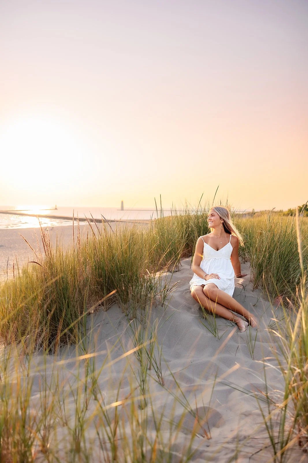 traverse city high school senior in a white sundress sitting on sandy beach with tall grass, enjoying the sunset with sailboats and a lighthouse in the distance.