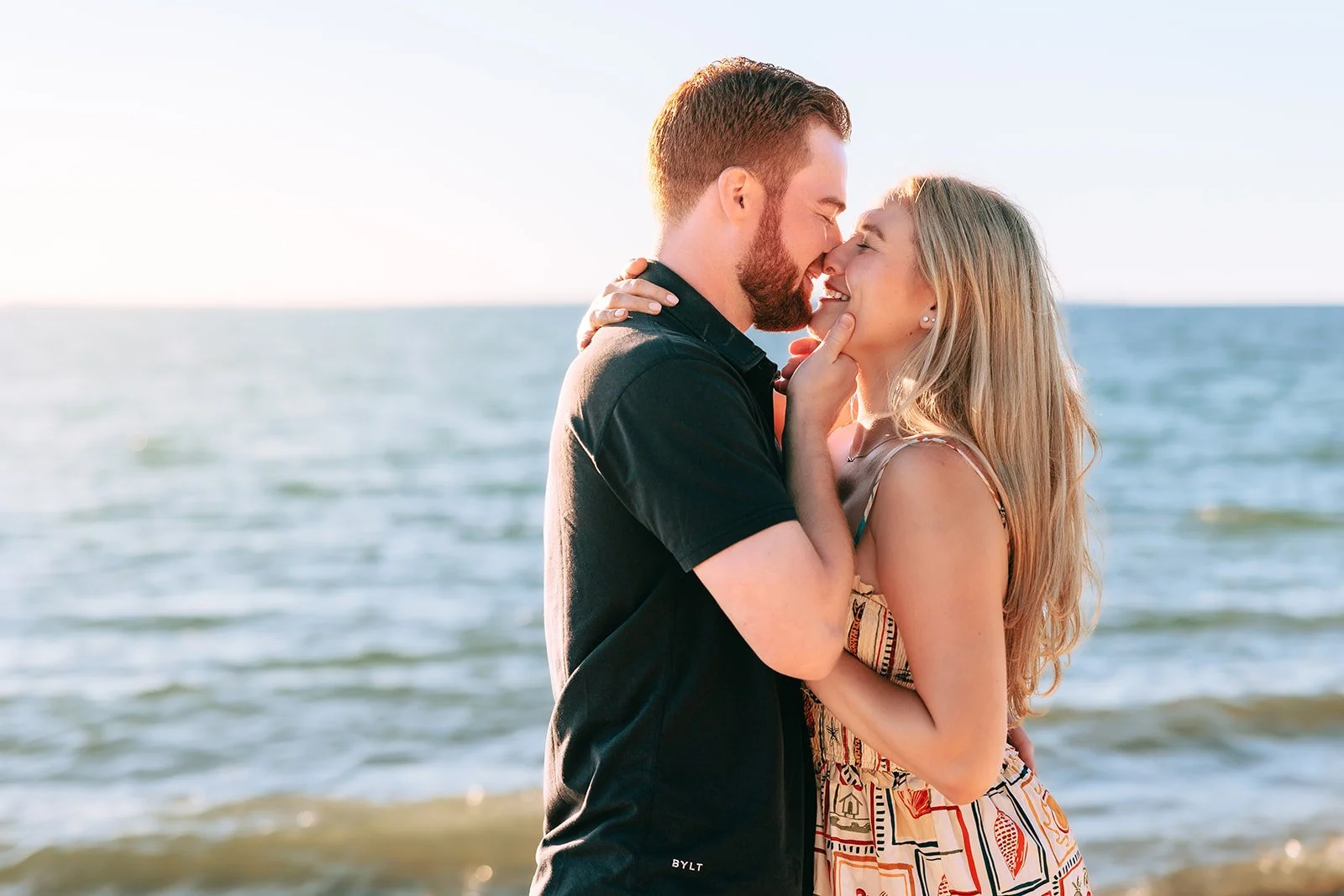 A loving couple standing close together by the water, touching noses and smiling, during sunset at the beach.