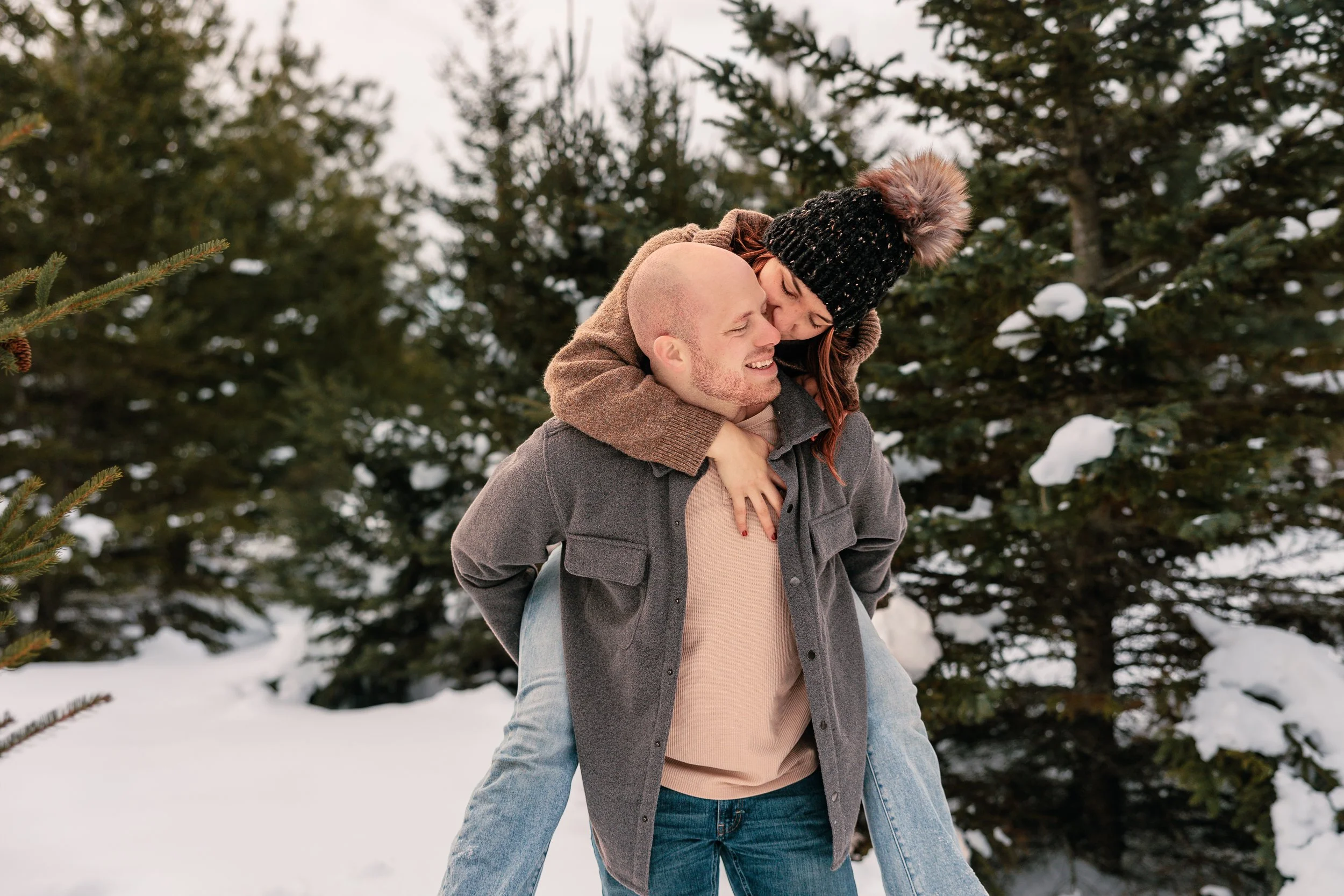 A couple enjoys a playful moment outdoors in a snowy forest, with the woman riding on the man's back as they smile and share an embrace.