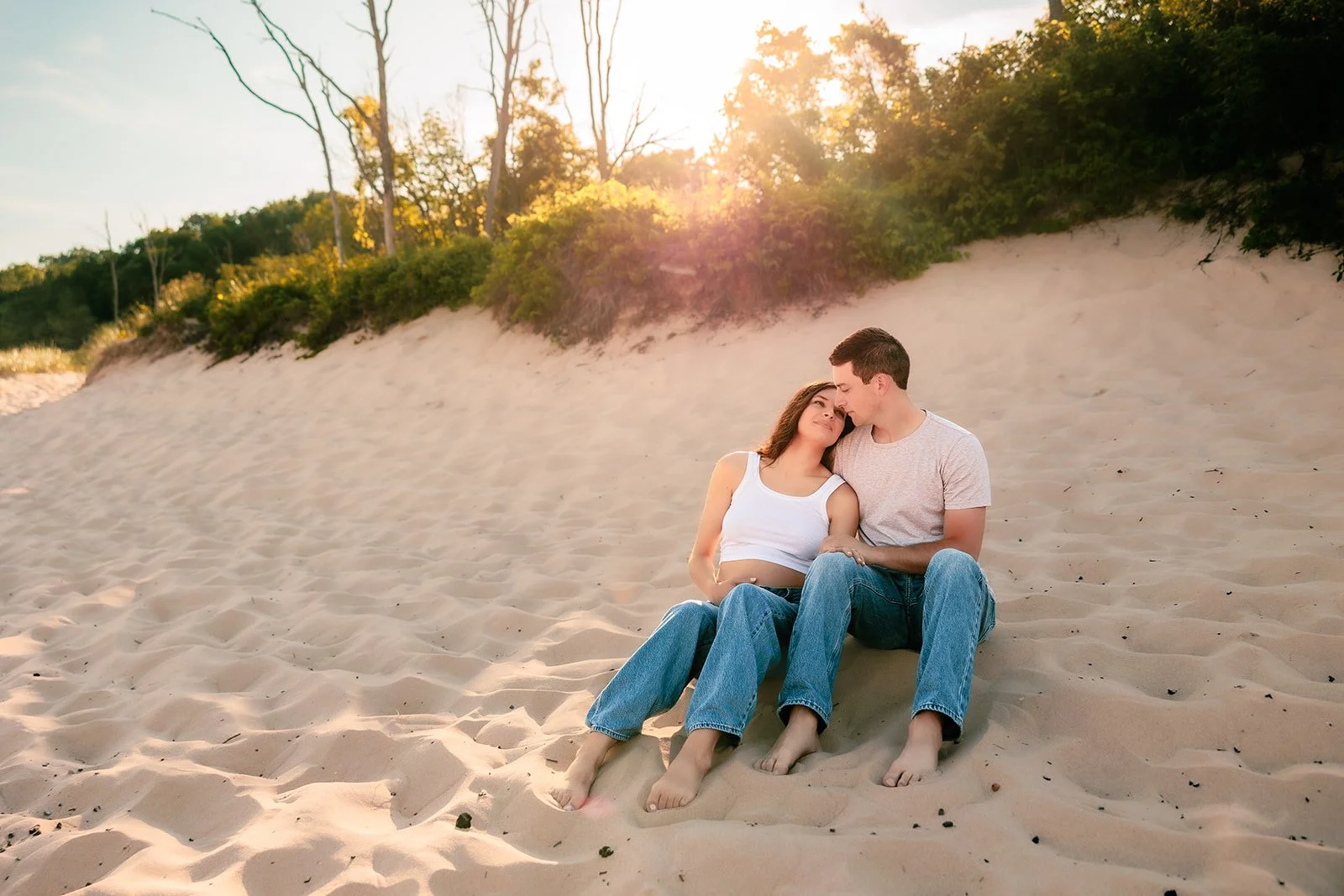 A couple sitting closely together on a sandy beach with green shrubs and trees in the background during sunset at esch beach looking over lake michigan