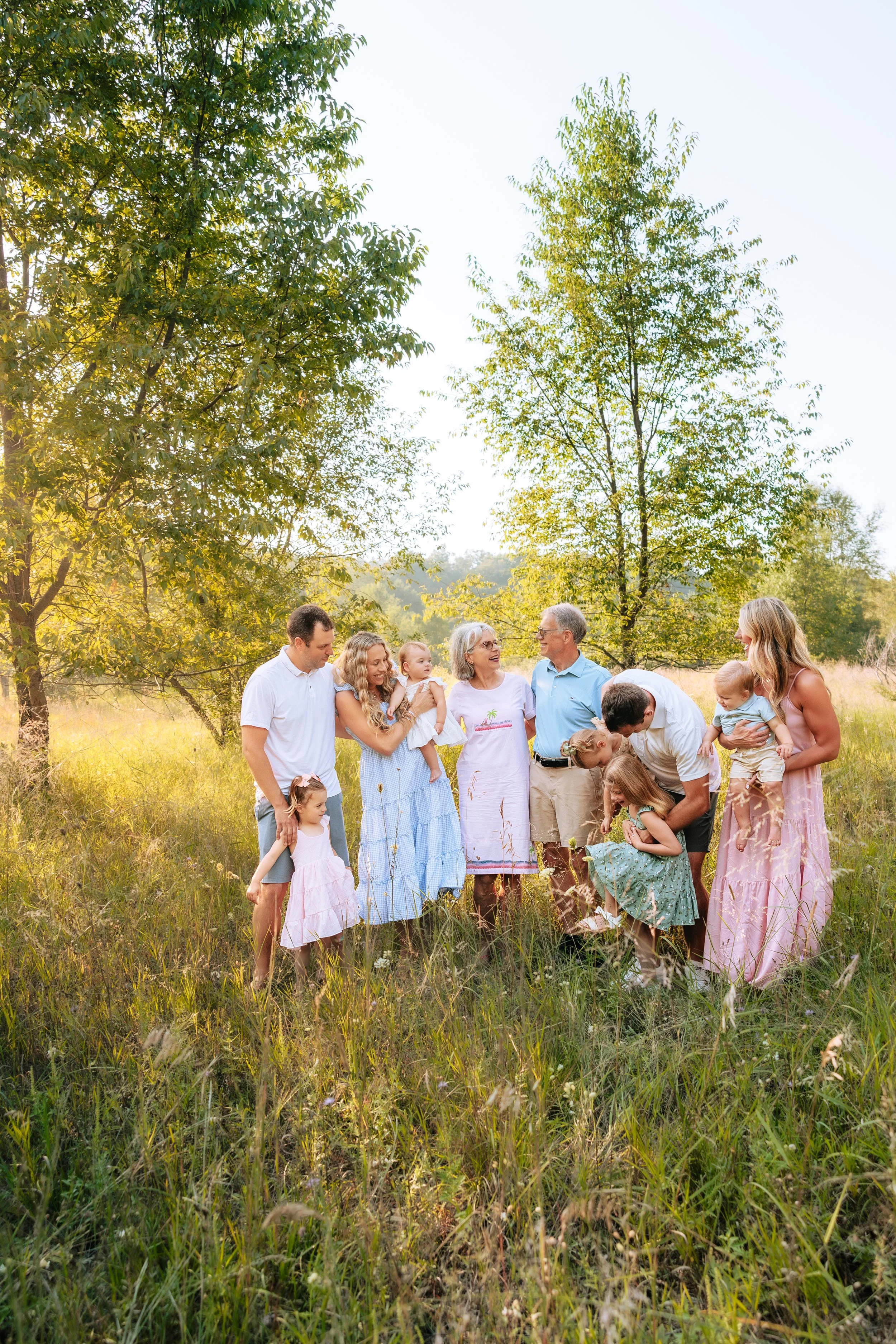 A multigenerational family of ten people, including children and elderly, gathering outdoors in a grassy field with trees, enjoying a sunny day.