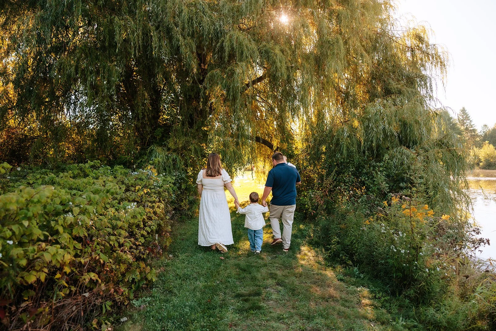 A family walking hand in hand along a grassy path near a pond in Leelanau during sunrise surrounded by lush green trees and foliage.