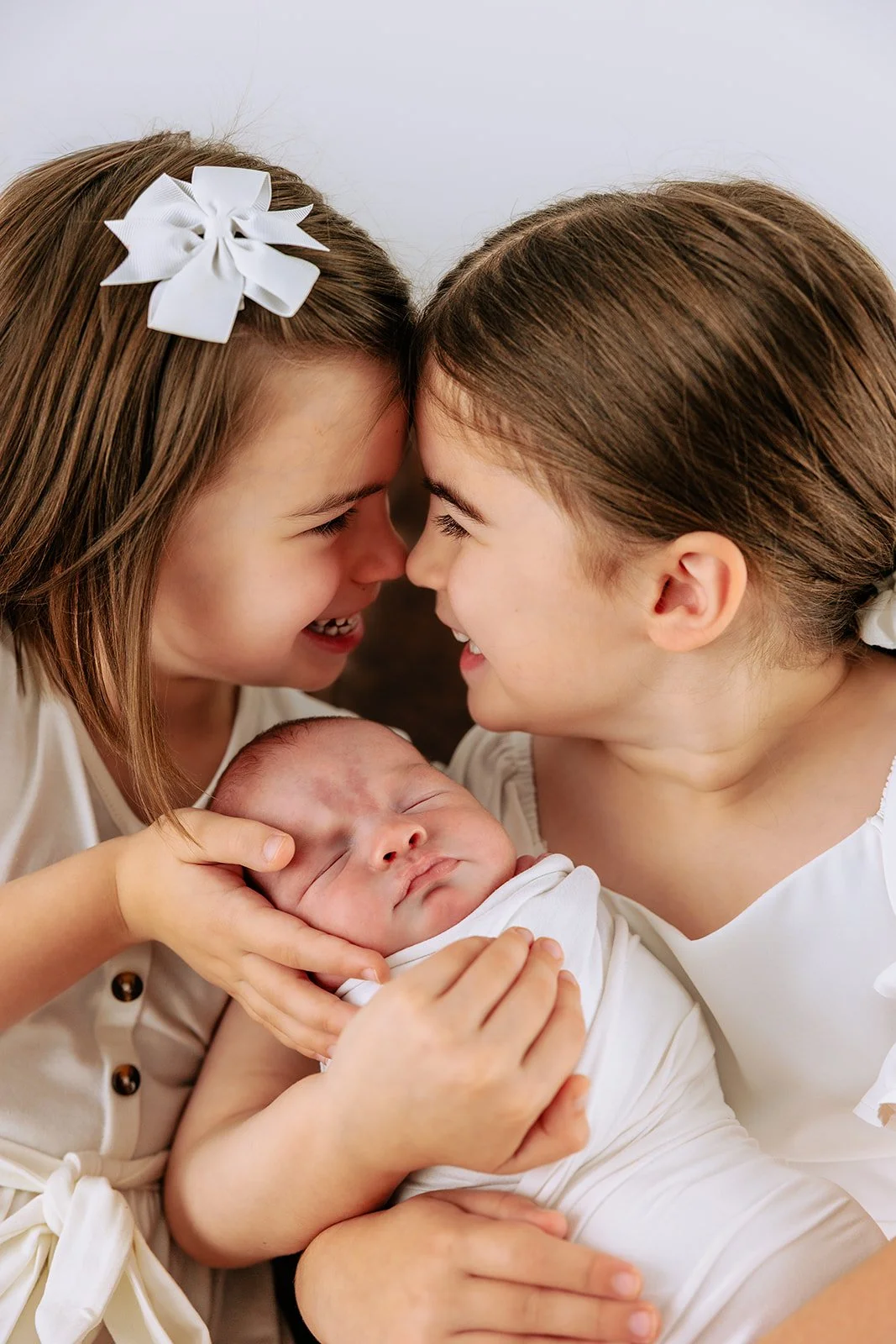 Two young girls, one with a white bow in her hair, smiling and touching foreheads, holding a sleeping baby with closed eyes, all dressed in light clothing against a plain background.