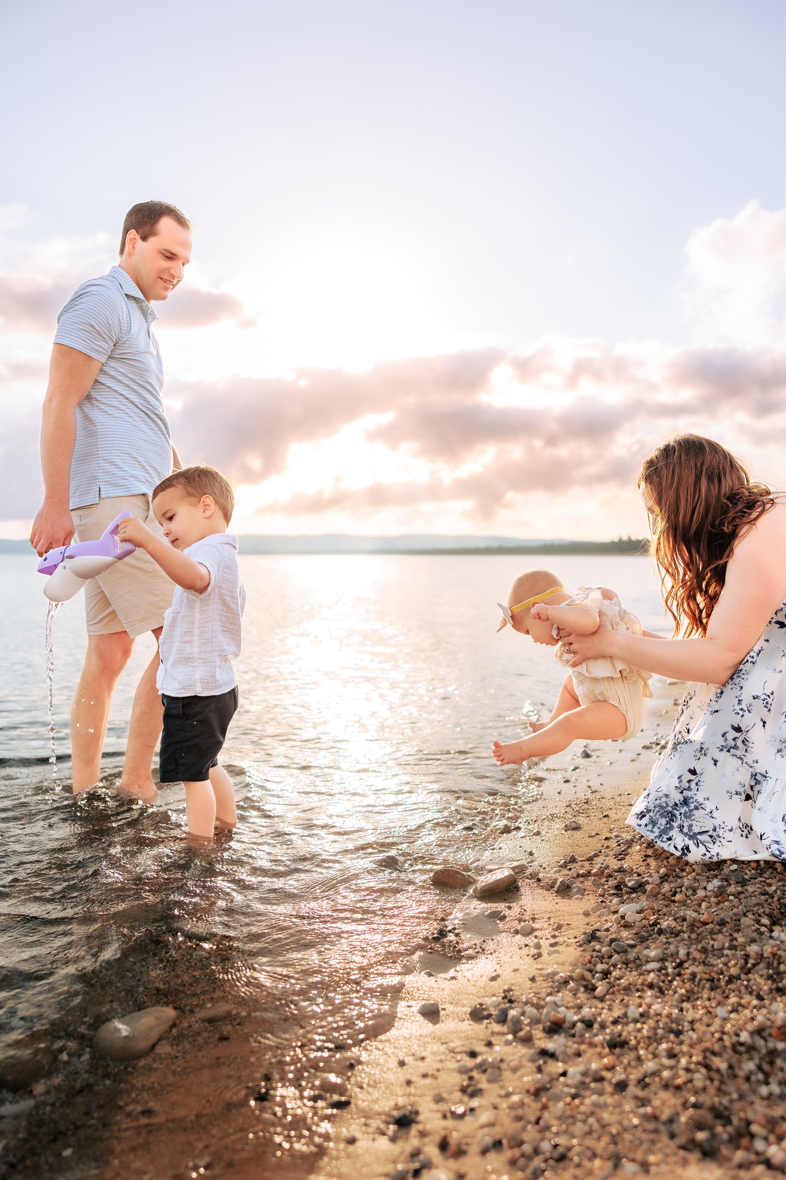 Family of four at the beach during sunset, with father and young son playing in the water, and mother and baby on the sandy shore.