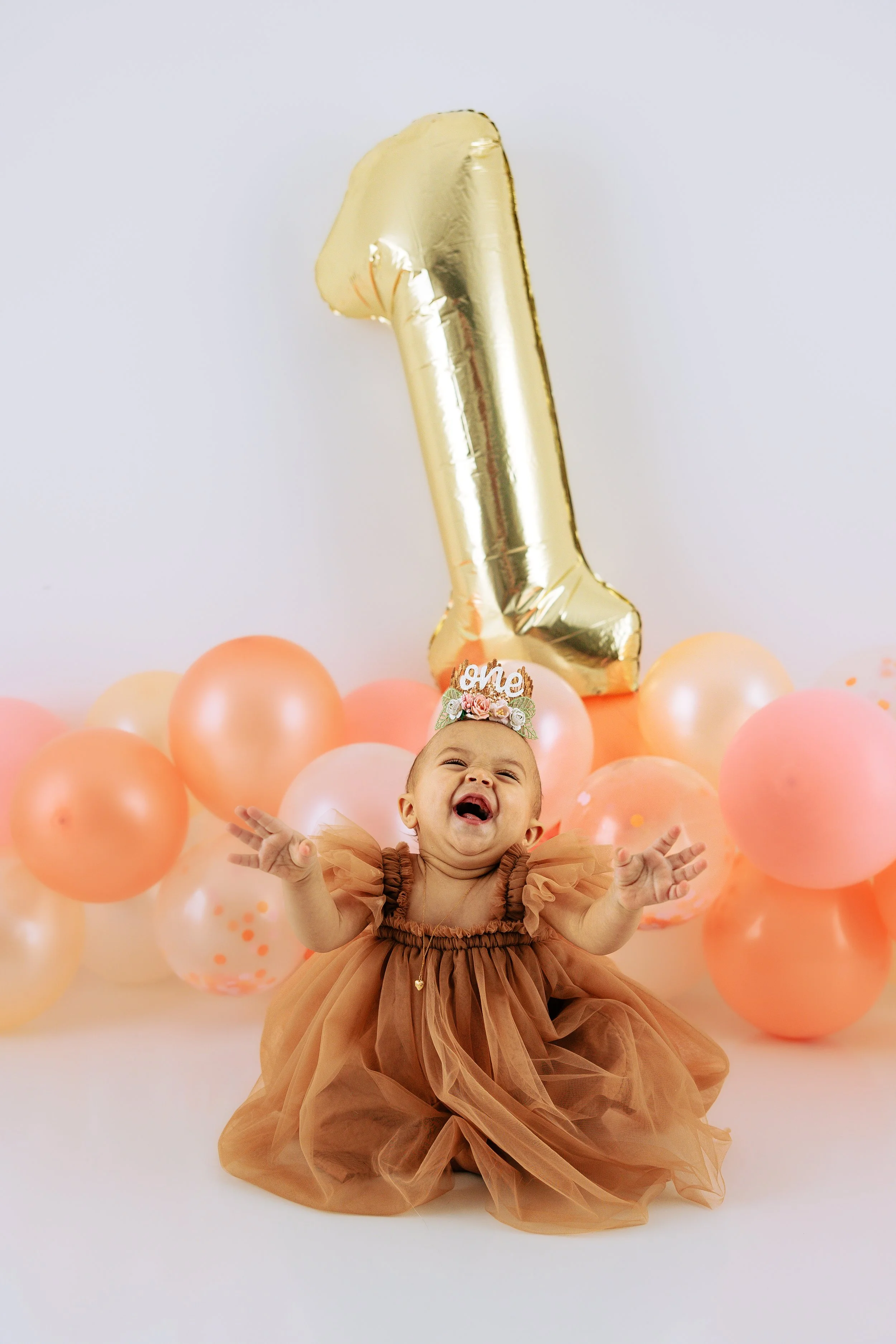 A baby girl in a brown dress celebrating her first birthday, sitting on the floor with a large gold number one balloon behind her, surrounded by peach and pink balloons, wearing a headband with a 'one' sign.