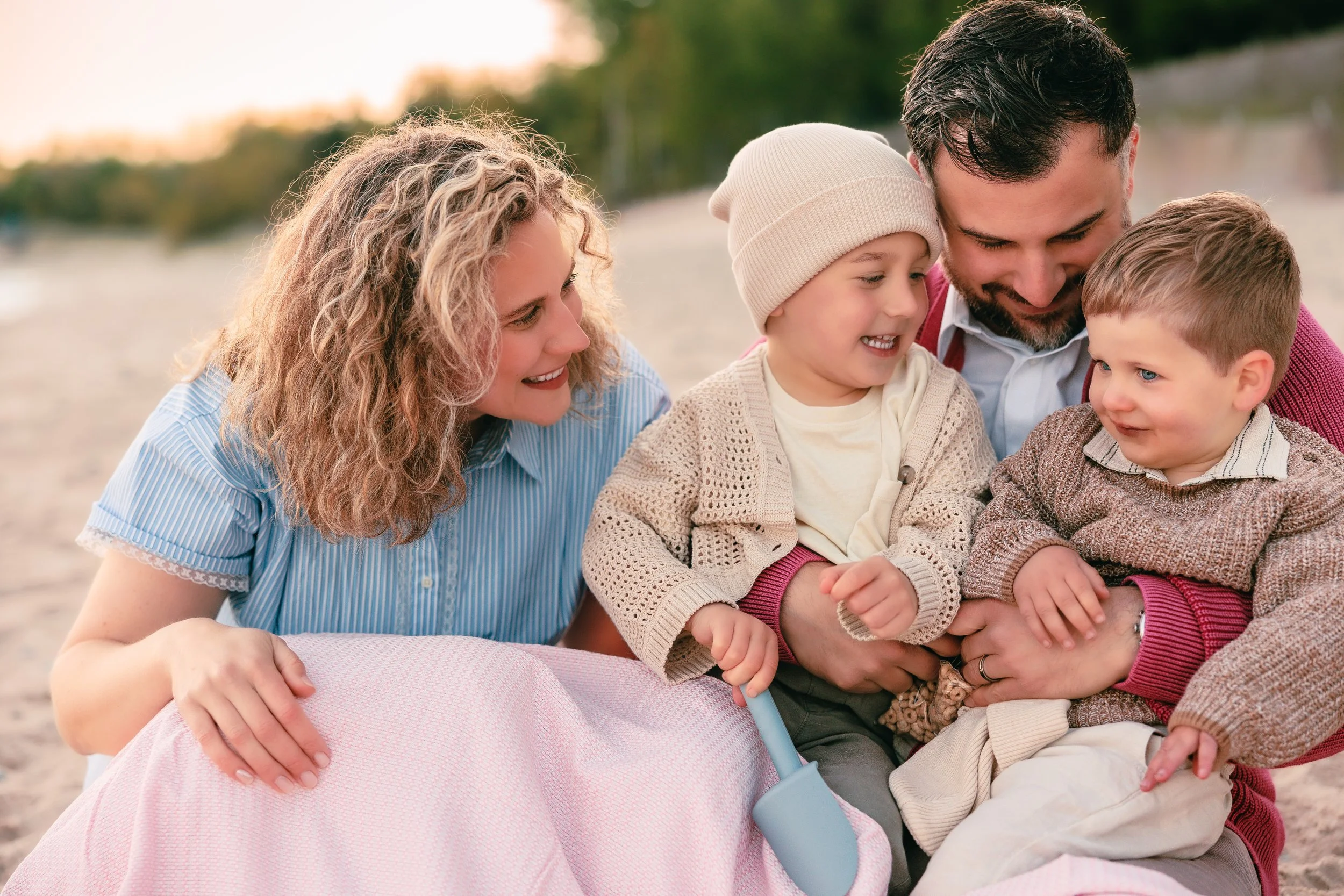 A family of four is sitting on a beach, smiling and playing together, with the lake and trees in the background during sunset.