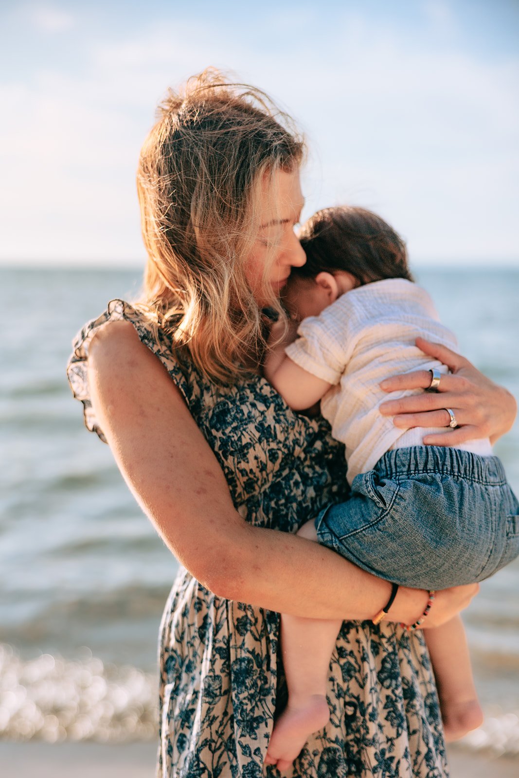 A woman holding a young child at the beach, with ocean and sky in the background. sleeping bear national park family session
