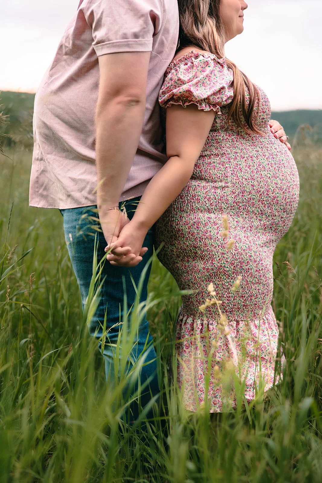 A pregnant woman and man holding hands in a grassy field, with the woman wearing a floral dress and the man wearing a light pink shirt and jeans, during sunset in sleeping bear national park