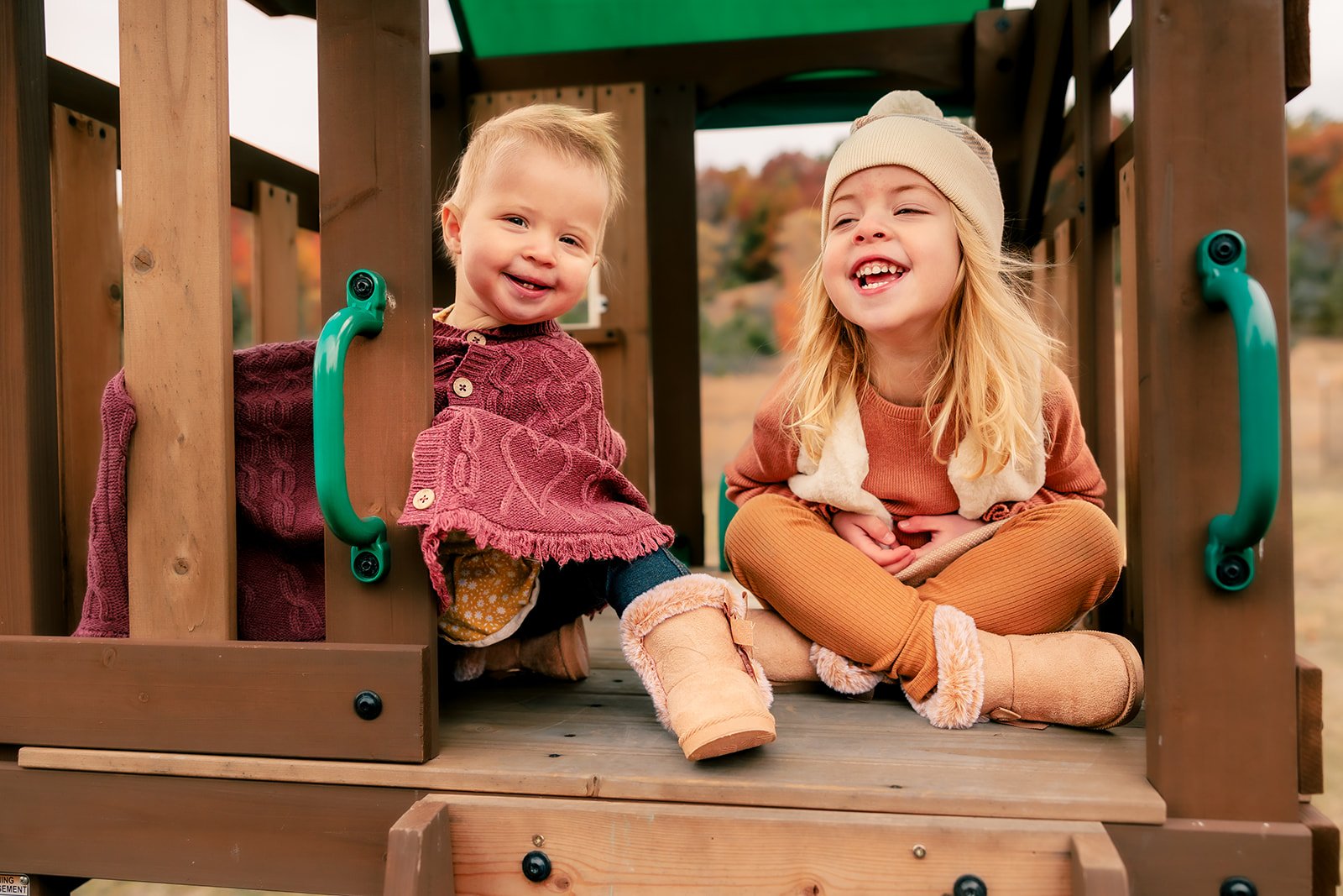 Two young children, a boy and a girl, sitting beneath a wooden playground structure outdoors during fall, dressed in warm clothes and smiling.
