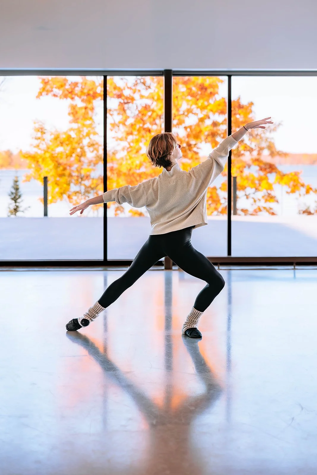 traverse city high school senior in dance attire practicing ballet or dance moves in a dance studio with large windows showing fall foliage outside.