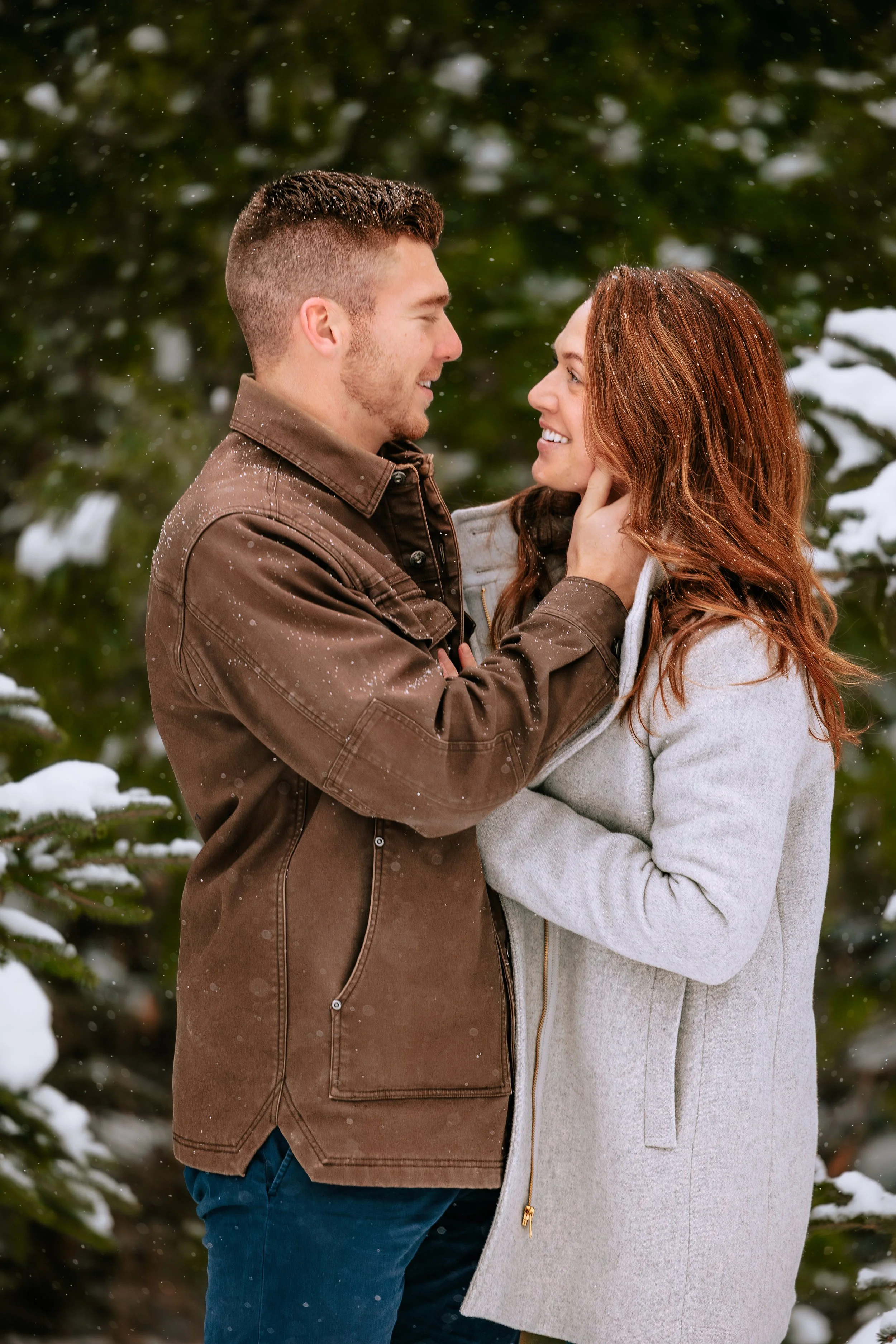 A couple standing close together in the snow, smiling and looking into each other's eyes. The man is holding the woman's face gently.