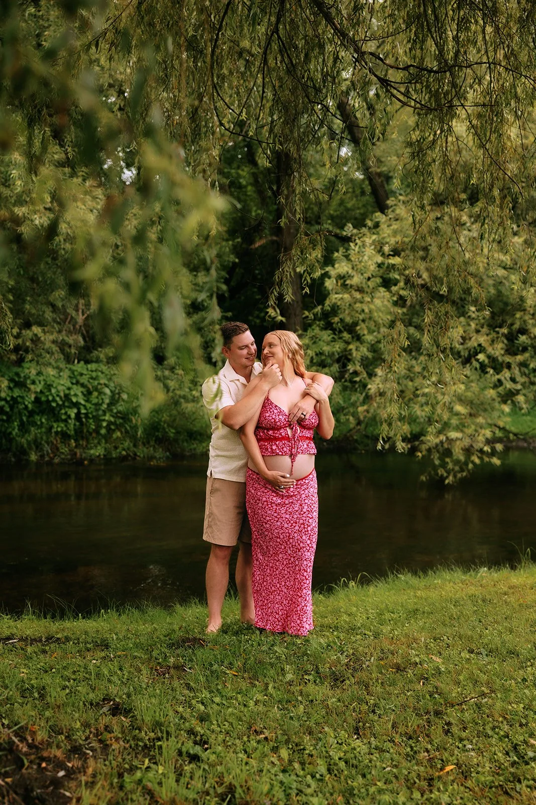 A couple stands by a river in a lush green park, the man holding a pregnant woman and gently touching her chin, both smiling lovingly at each other in northern michigan