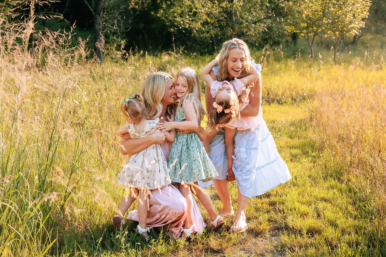 A group of two women and three young girls laughing and hugging in a grassy field during golden hour with trees in the background northern michigan family