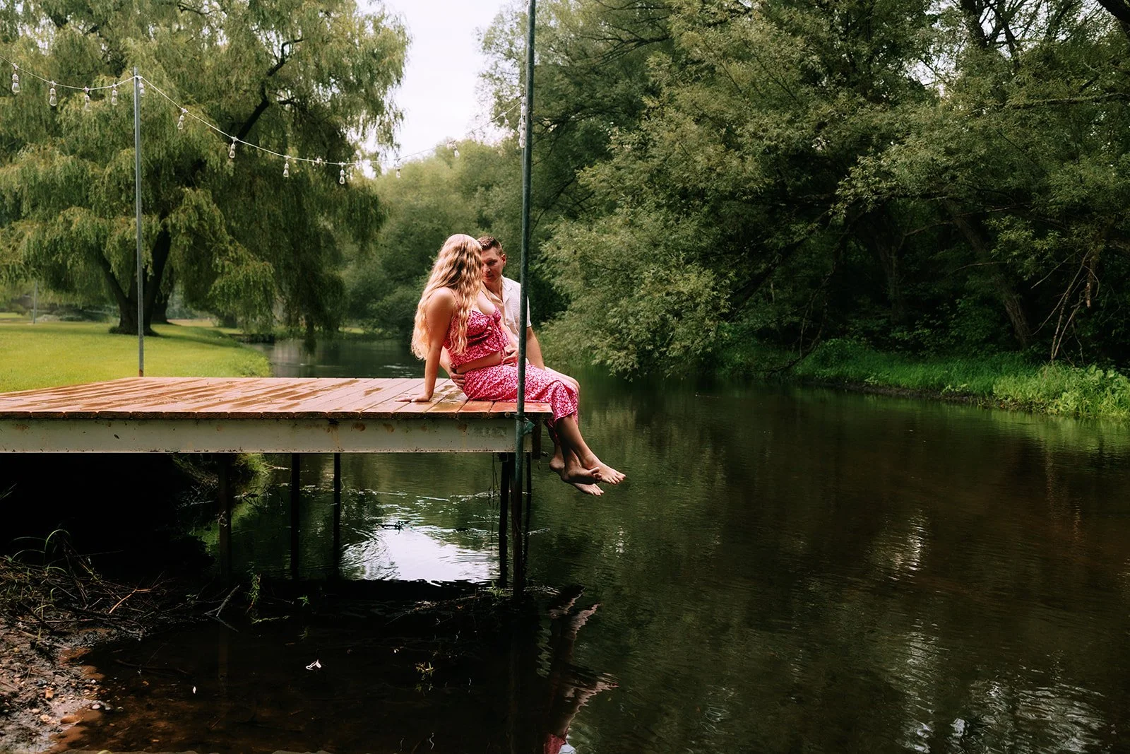 A young couple sits on the edge of a wooden dock over a calm river in a lush, green park, engaging in conversation in northern michigan