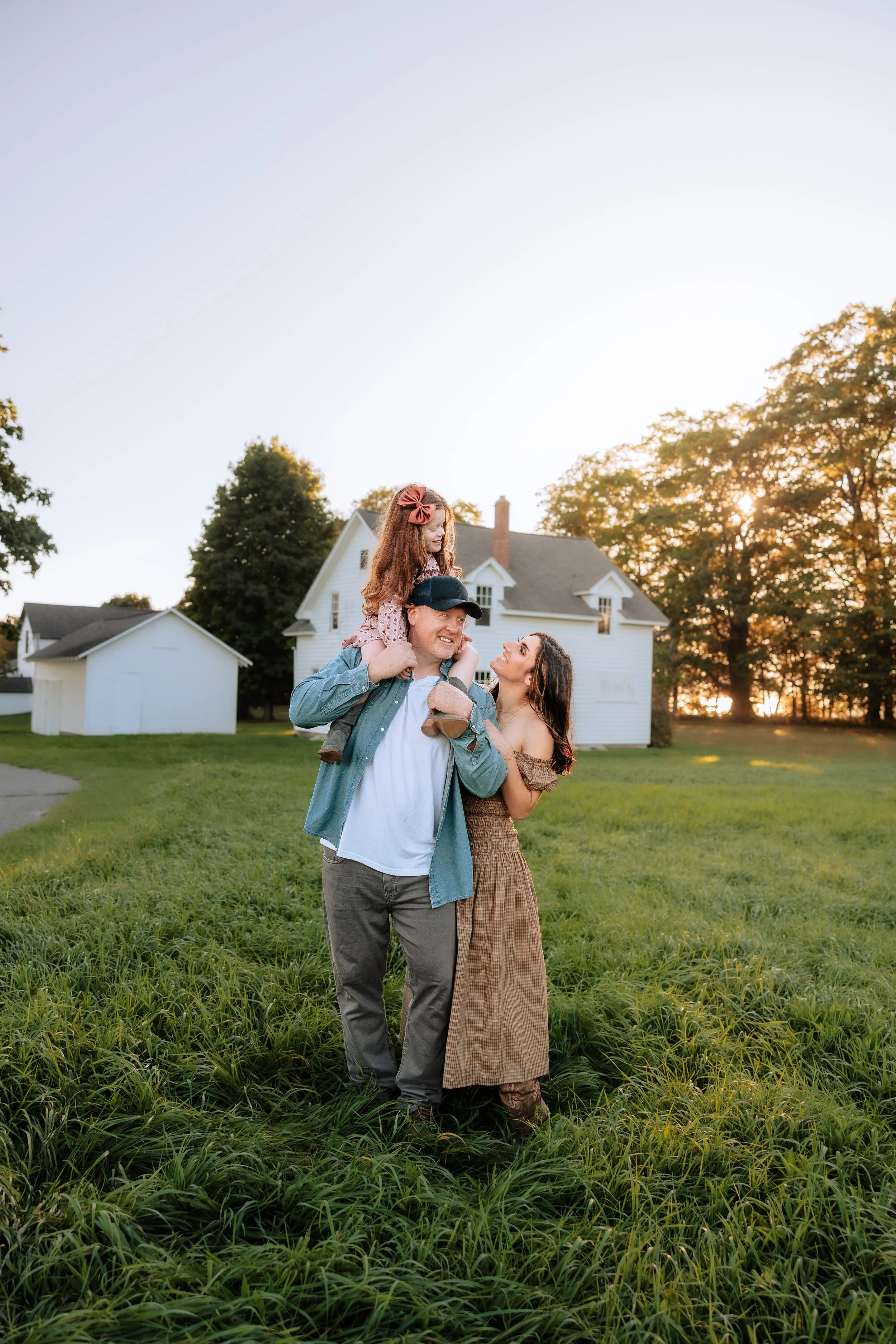 A family of three, with a man carrying a young girl on his shoulders and a woman standing next to him, walking through a grassy field at sunset in front of a white house and trees.