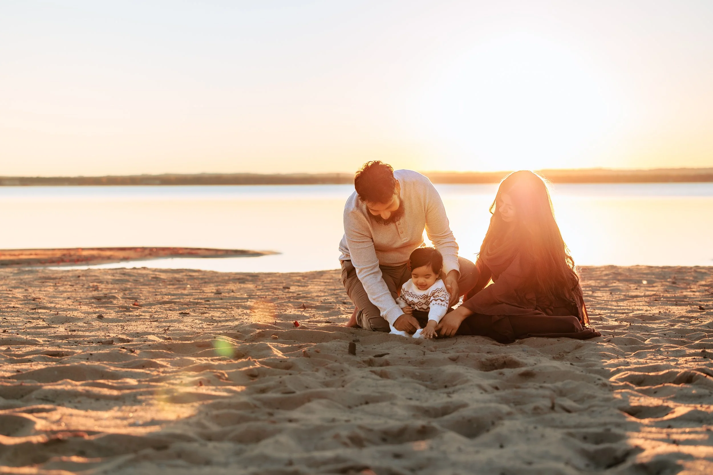 A family of three sitting on a sandy beach at sunset, smiling and playing with their young child.