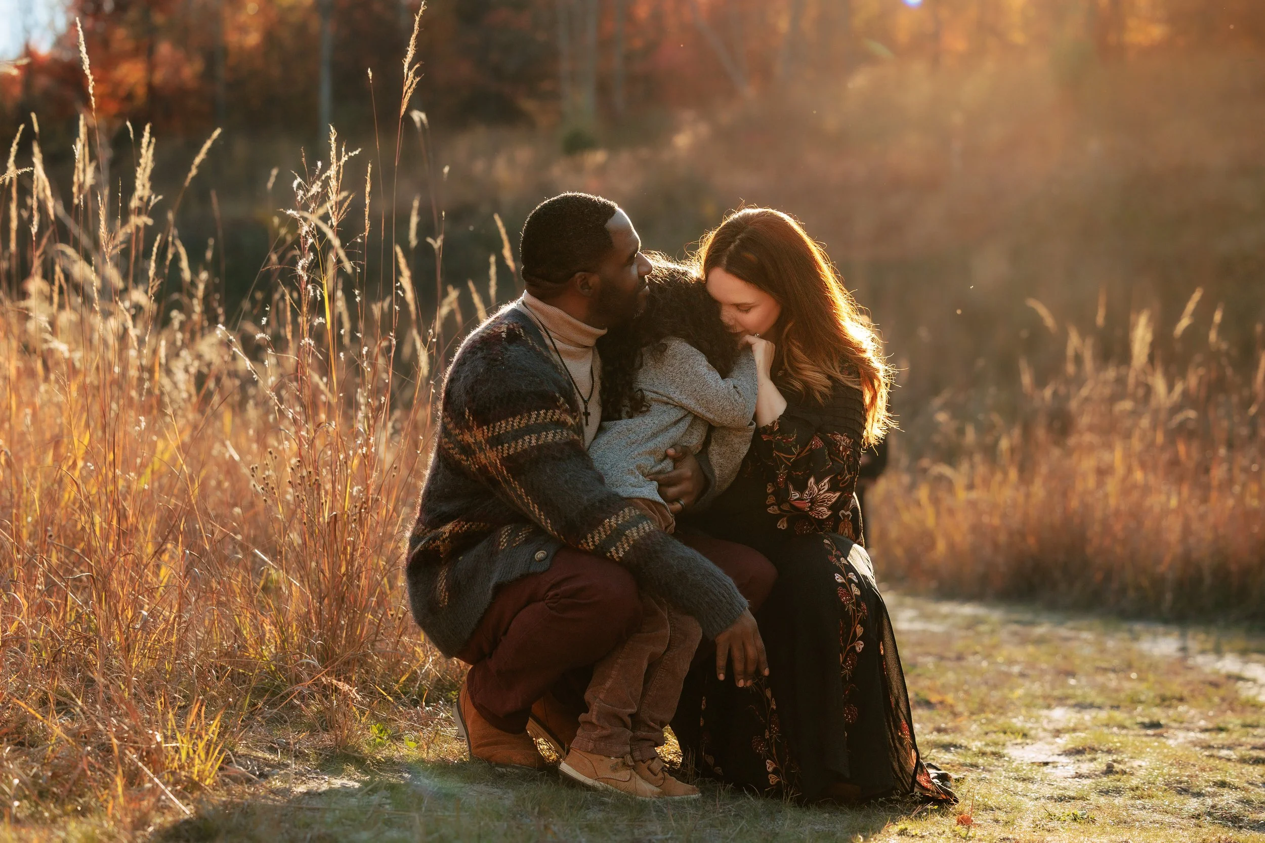 A family of three, a man, woman, and young girl, sitting outdoors in a field with tall grass and autumn trees in the background, during sunset, sharing a tender moment.