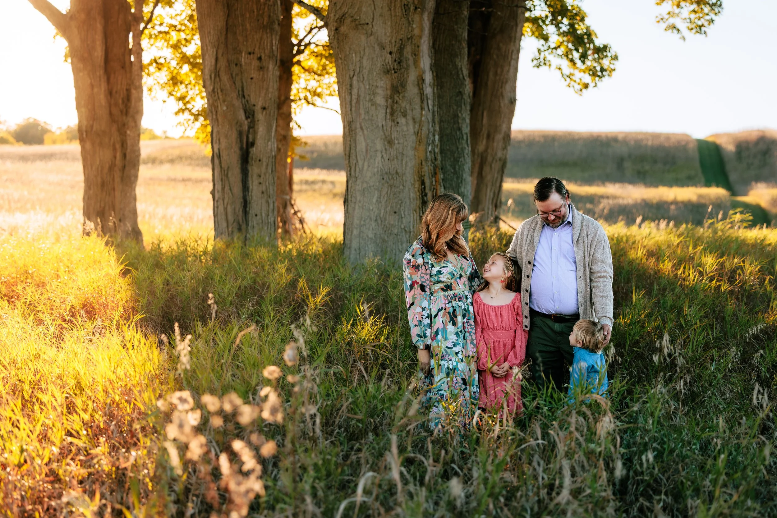 A family of four, including two adults and two children, stands together in a lush, grassy field with tall trees during the golden hour of sunset.