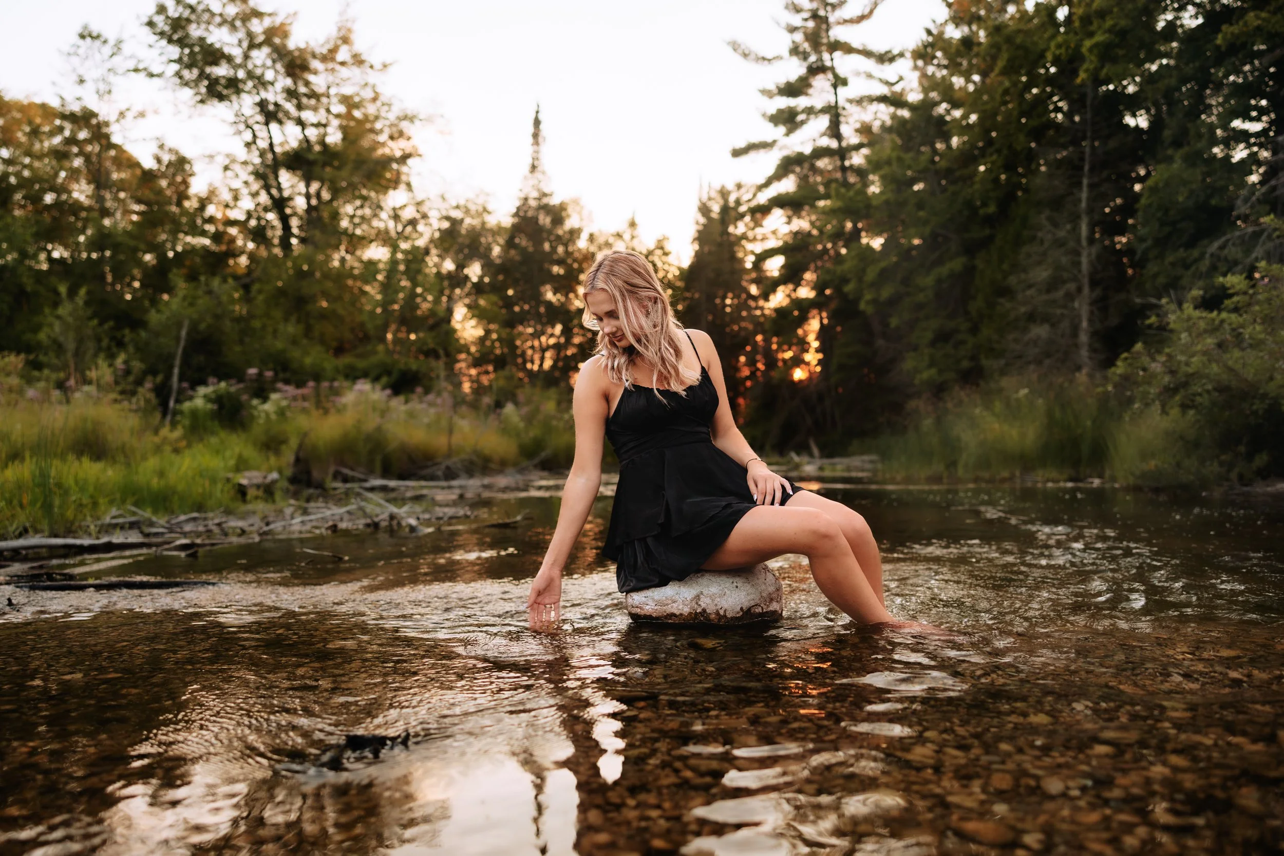 A woman in a black dress sitting on a rock in a shallow river, touching the water during sunset in a forested area.