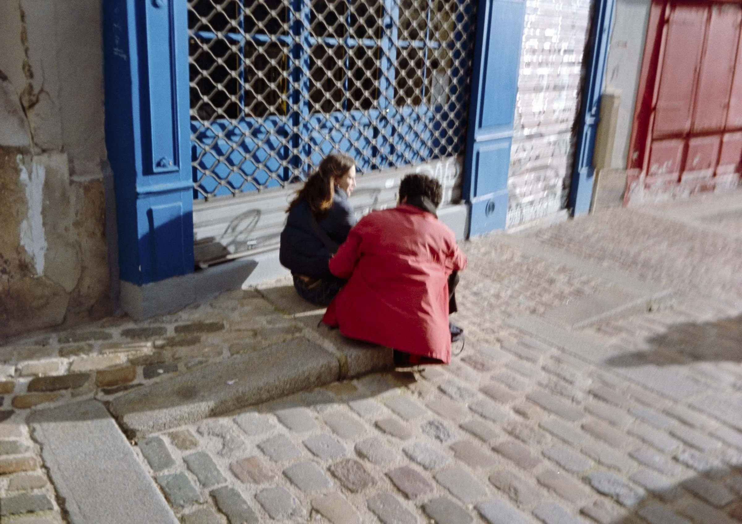 Two people sitting on a curb outside a building with blue and red shutters, engaged in a conversation.