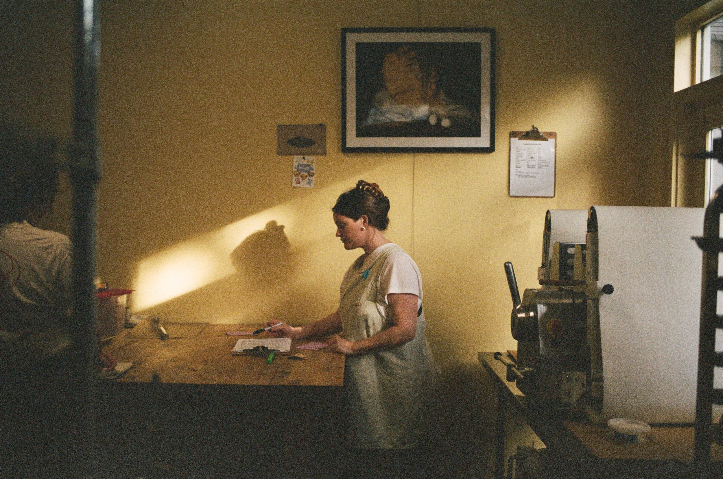 A woman in a white t-shirt and apron standing at a wooden counter in a yellow-walled room, holding a pen and looking down, with a framed picture hanging on the wall behind her.