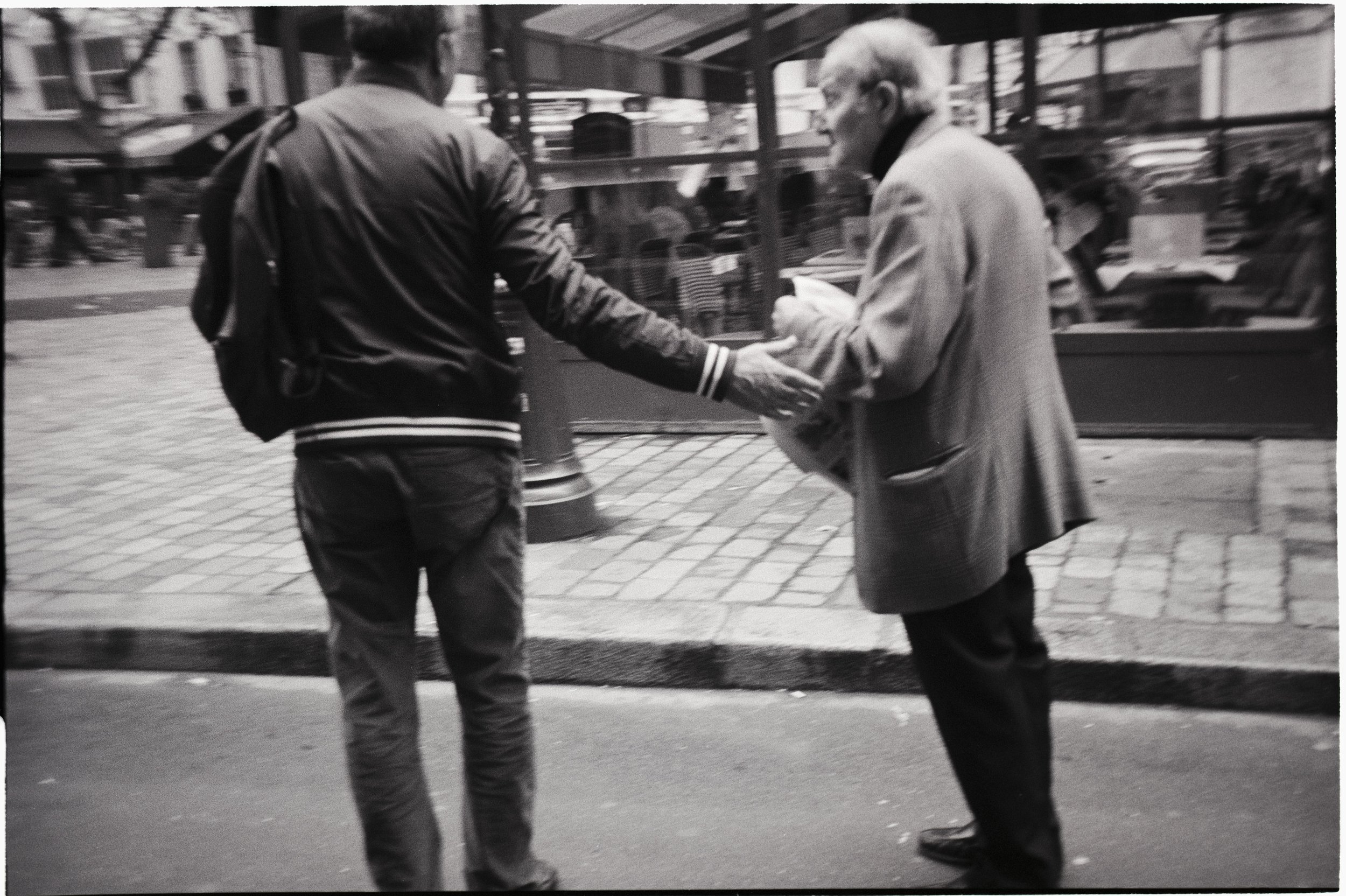 A black and white photo of two people standing on a city sidewalk, one person is handing something to the other. The person on the left is wearing a jacket with a backpack, and the person on the right is an older woman with short light-colored hair, 