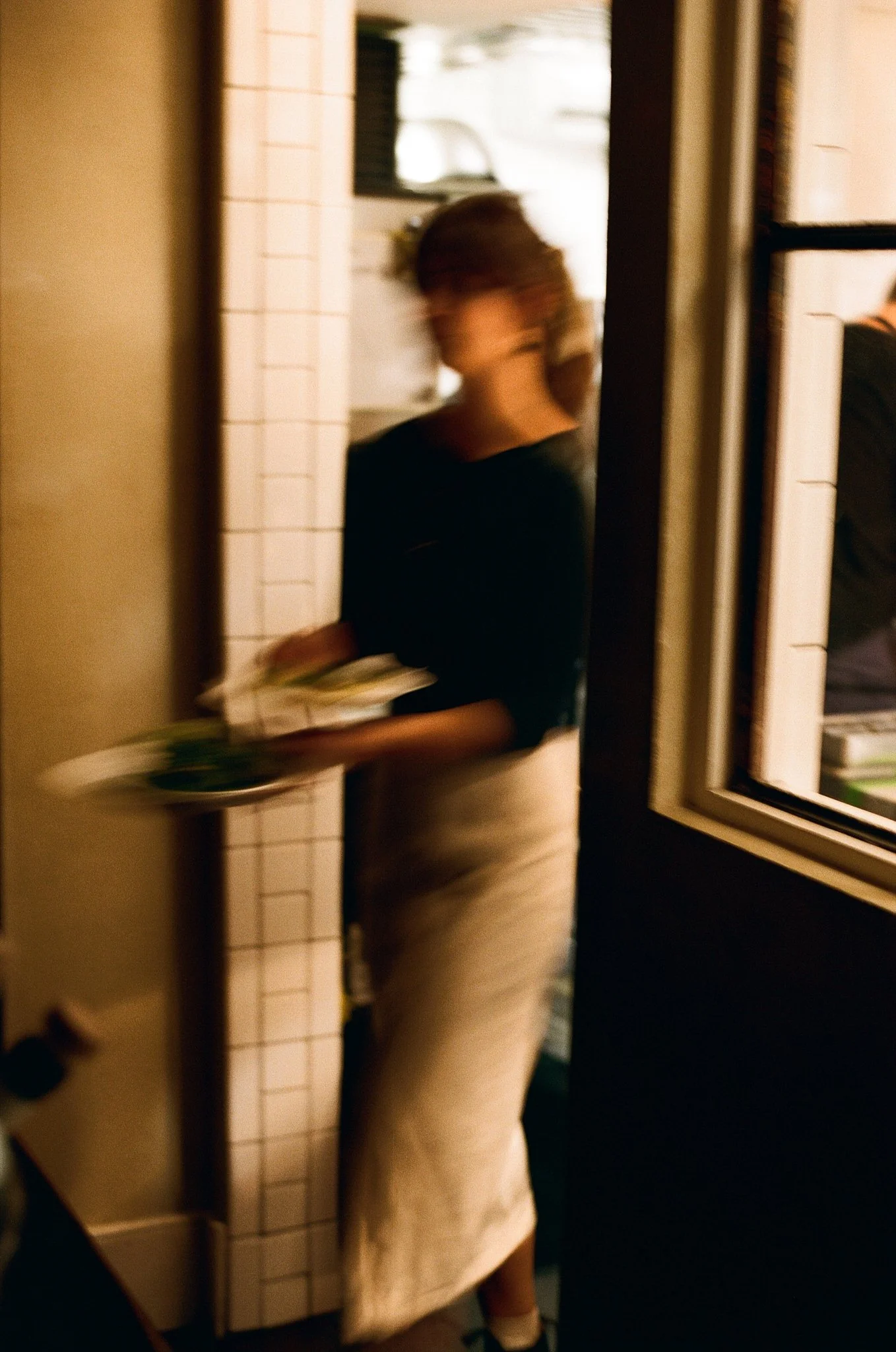 Woman with short hair, wearing a black top and beige skirt, walking through a doorway holding a tray of food.
