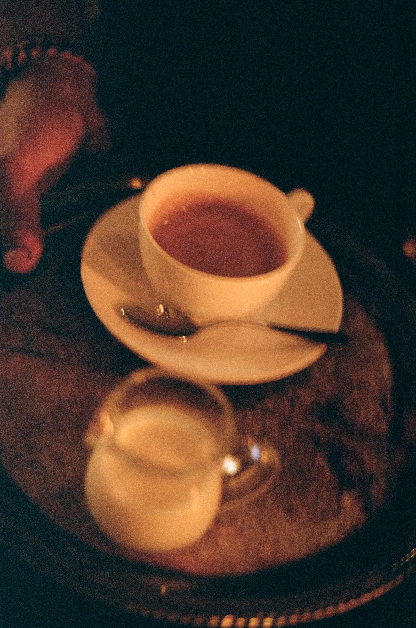 A white cup of hot tea on a saucer with a small spoon, and a small white pitcher, on a dark surface, in a dimly lit setting.