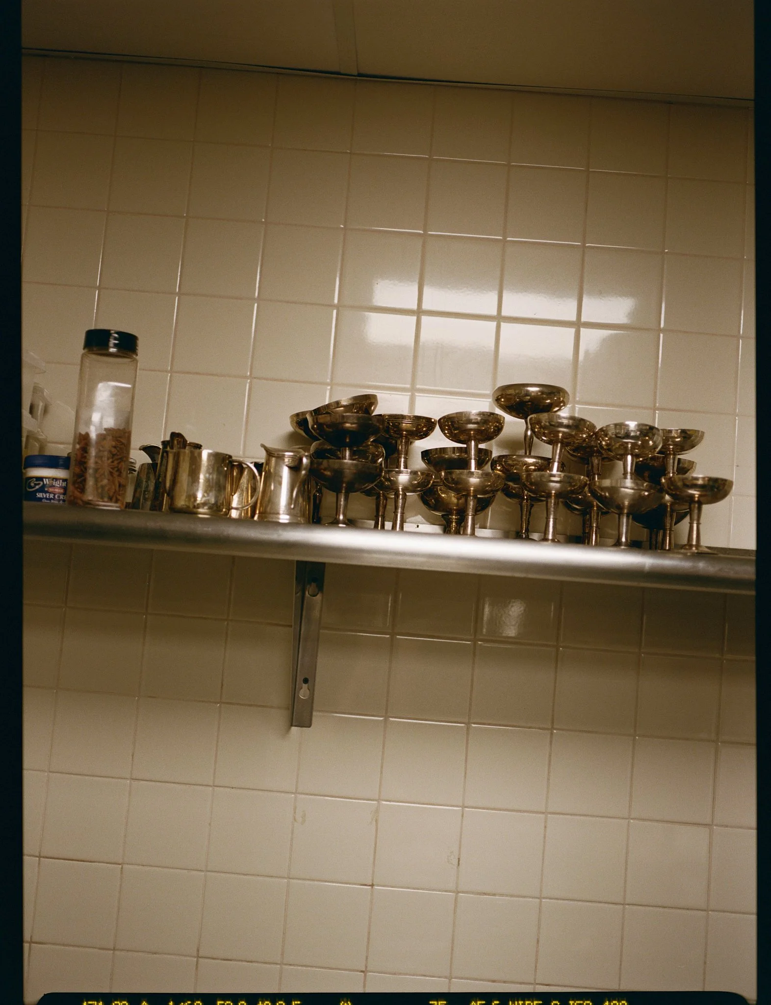 A kitchen shelf with a row of silver dessert cups and small metal pitchers against a tiled wall.