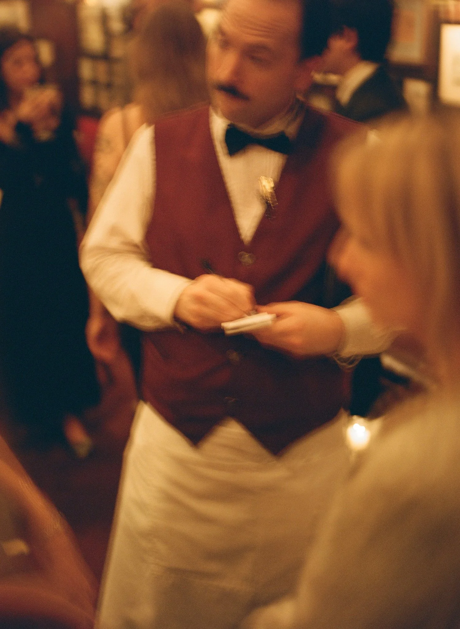 A waiter dressed in a red vest, white shirt, and black bow tie taking notes while talking to a guest at a busy restaurant.