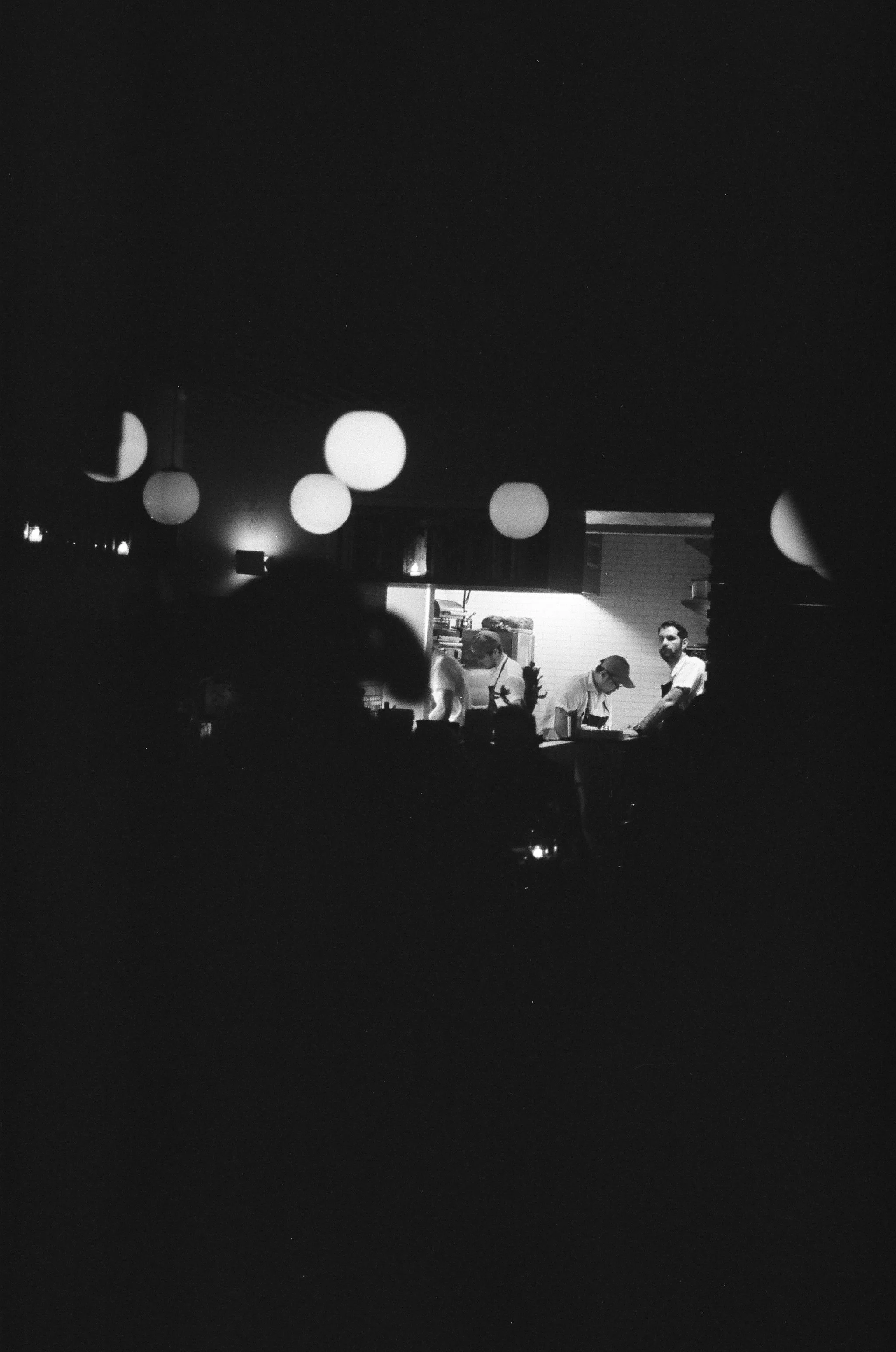 Black and white photo of a kitchen seen through a window with hanging lanterns, featuring four people working or preparing food.