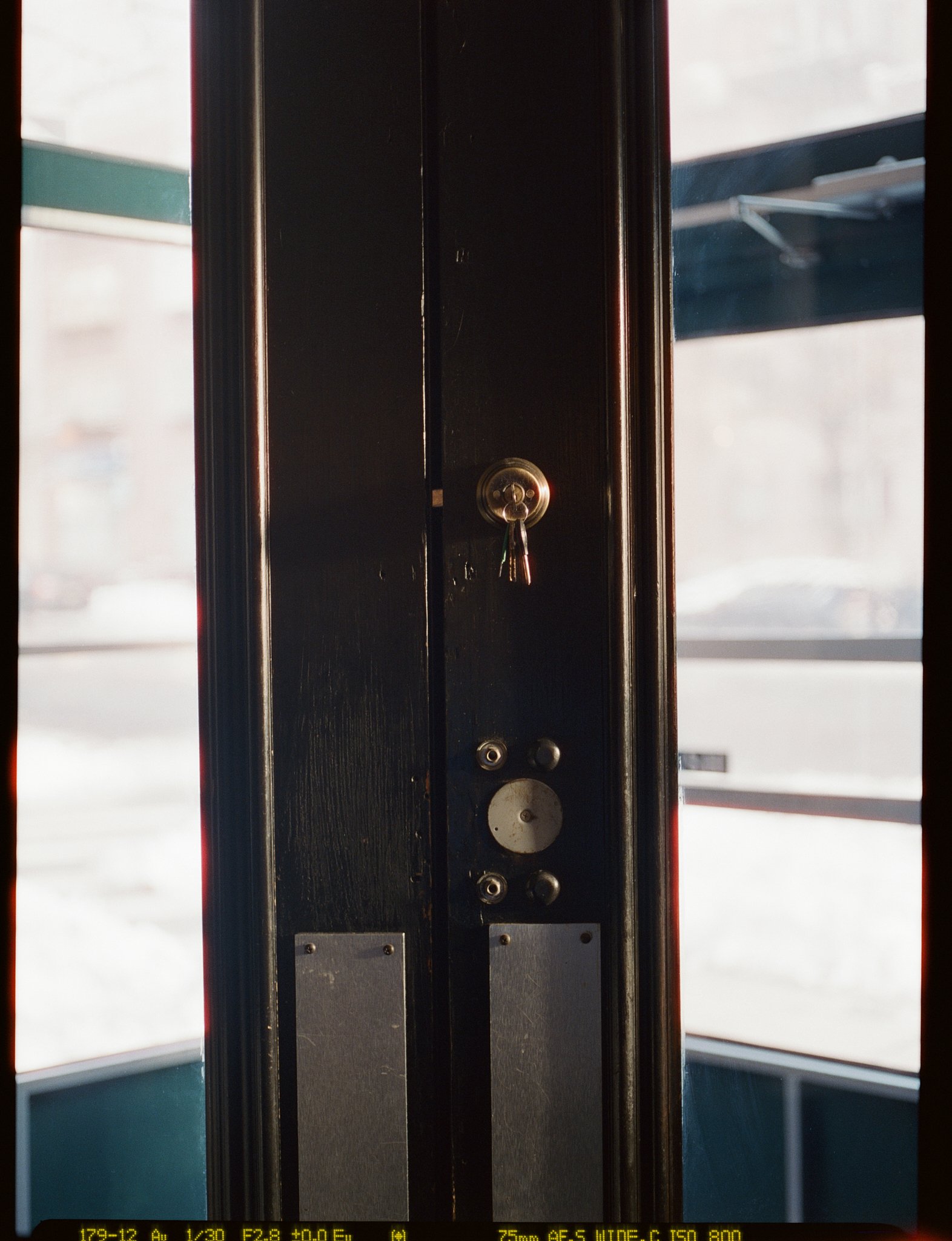 Close-up of a door frame with a brass lock and no handle, seen from inside a building with large windows, showing an outside street scene.