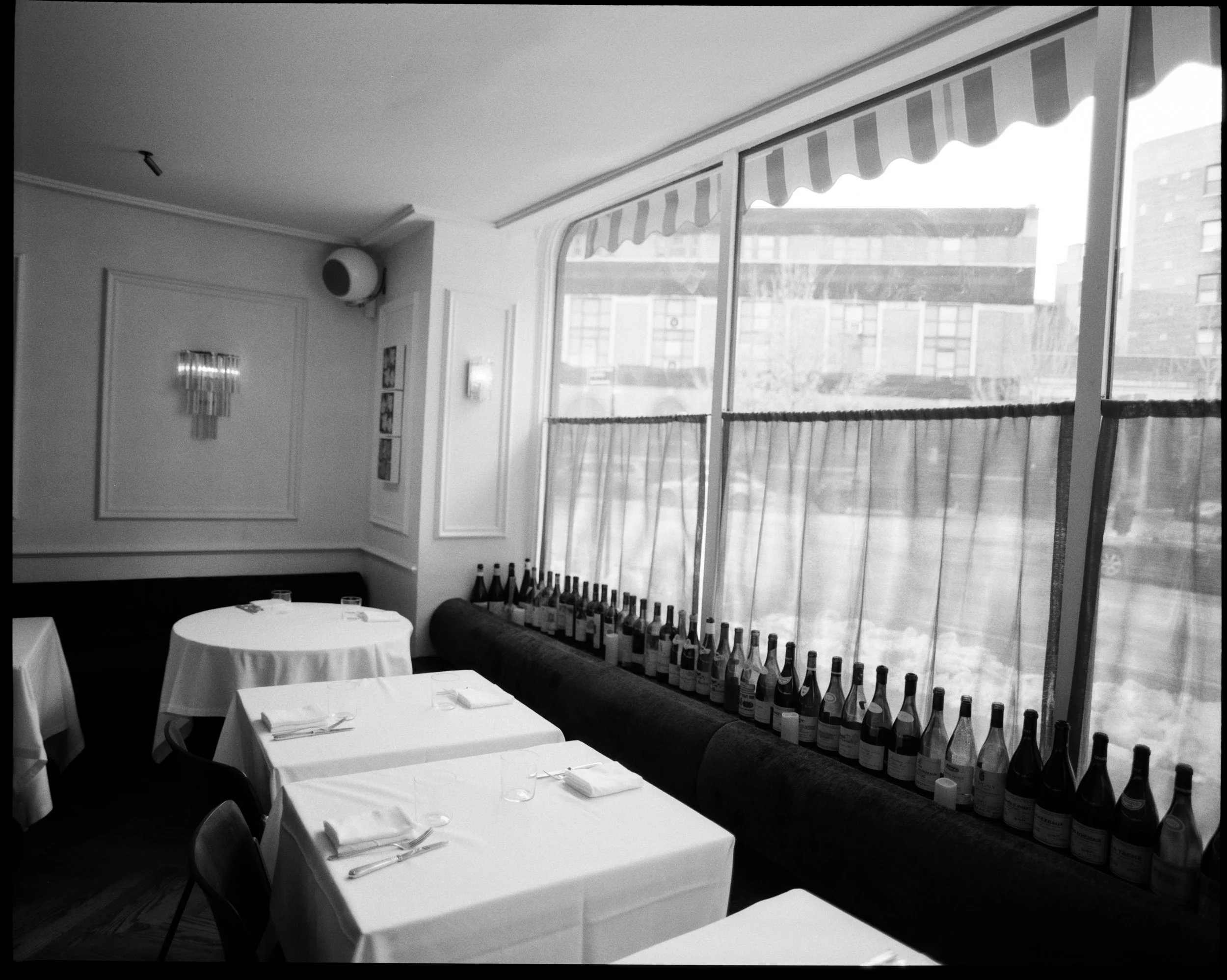 An empty restaurant dining area with white-draped tables set with silverware and napkins, a row of wine bottles along a cushioned window seat, and large windows with striped awnings showing a cityscape outside.