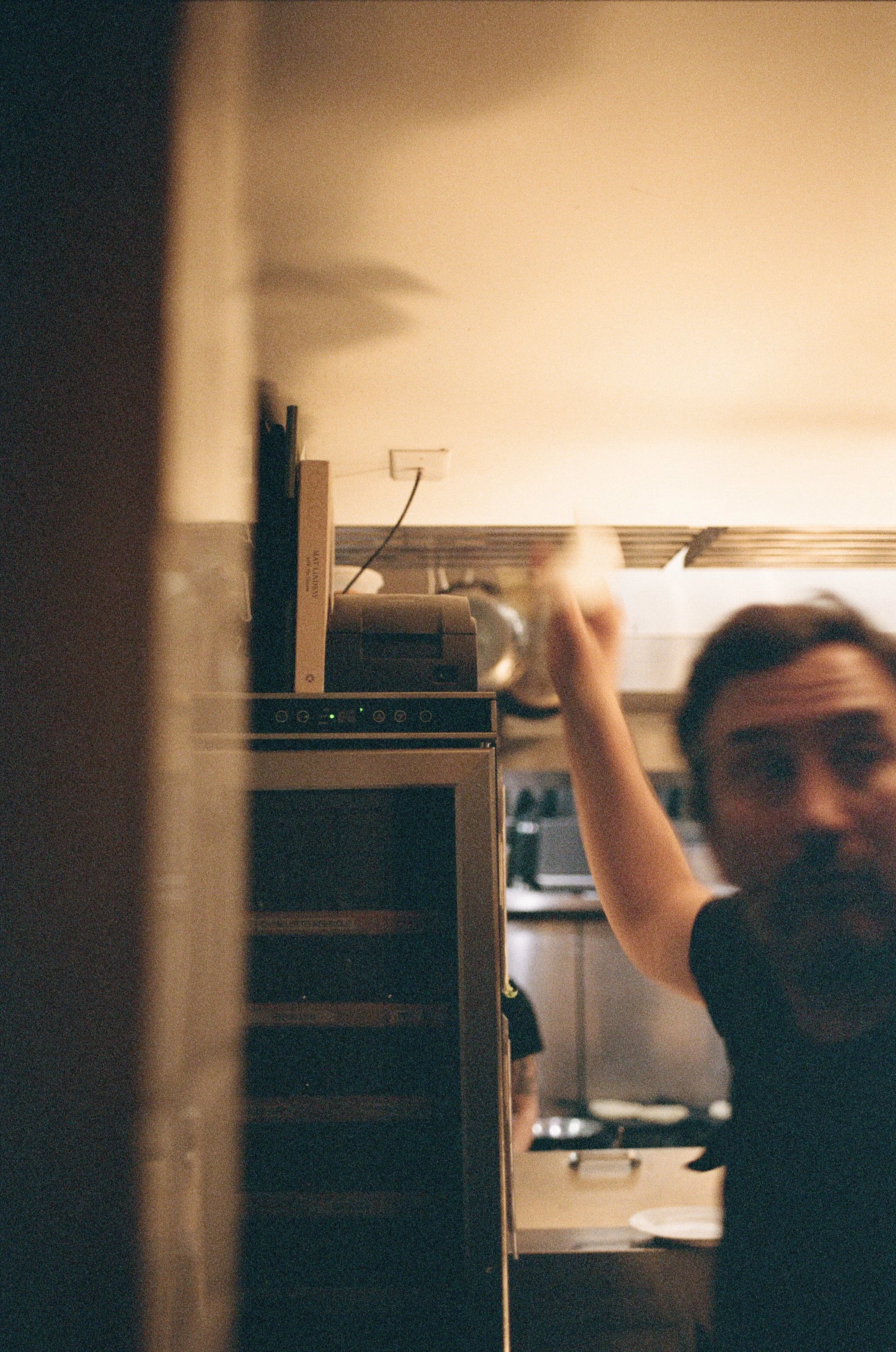 A person with glasses and a beard in a kitchen, reaching towards the fridge or shelf.