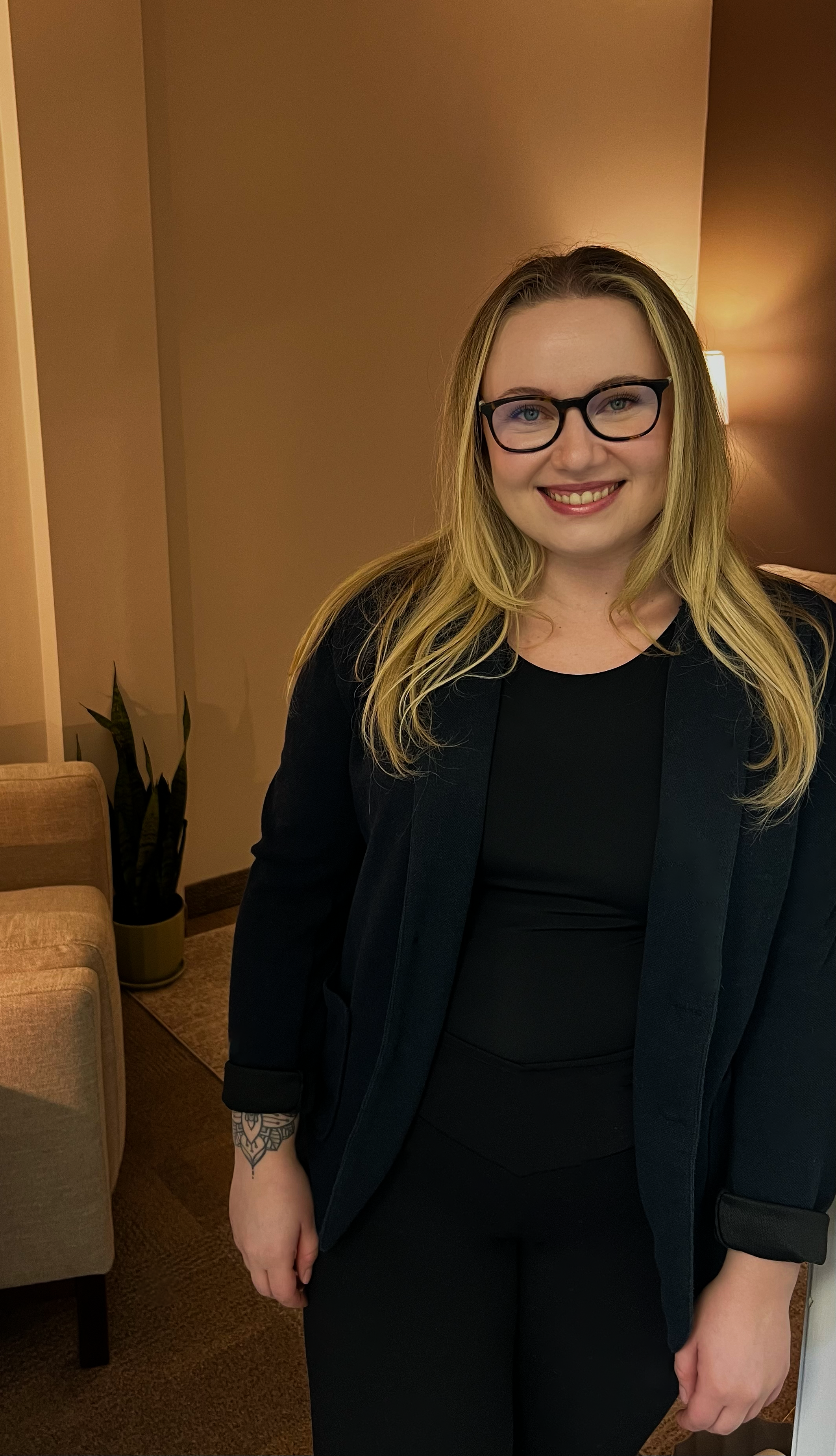 A woman with blonde hair, black glasses, and a tattoo on her wrist, wearing a black top and blazer, smiling indoors with warm lighting.
