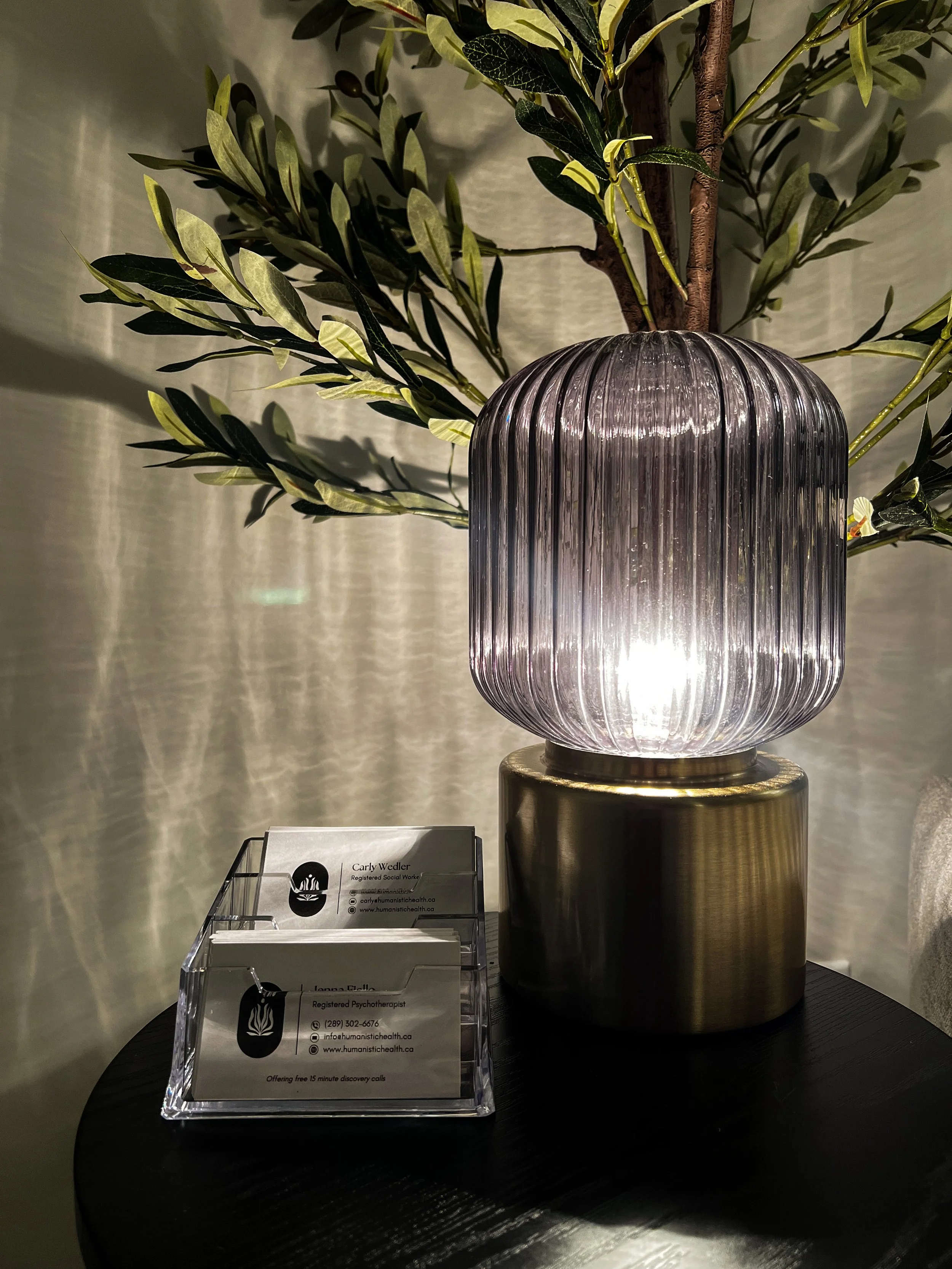 A decorative table lamp with a ribbed glass shade and a brass base, placed on a round black table alongside business cards in a clear holder. A leafy plant is in the background.