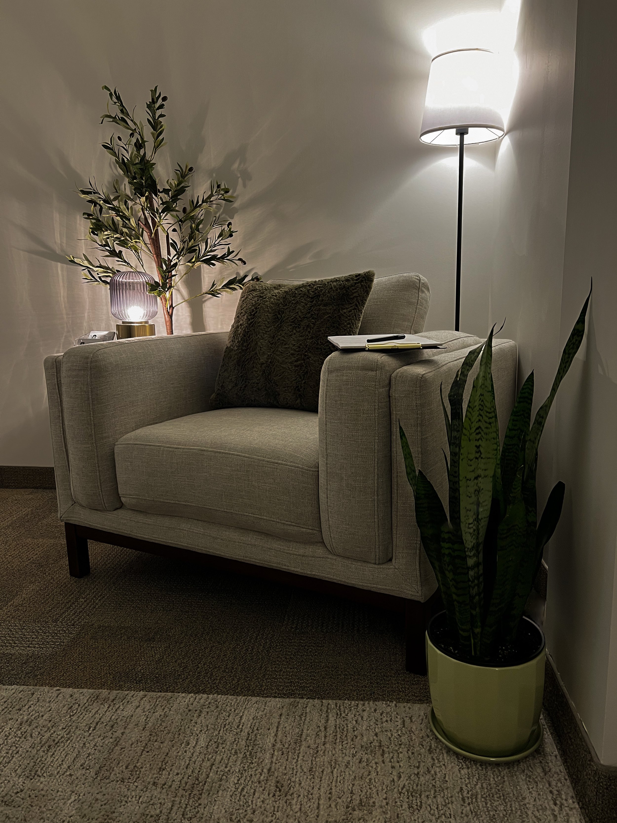 Comfortable beige armchair with a dark green pillow, a small notebook and pen resting on the arm, flanked by tall indoor plants, with a standing lamp and a decorative table lamp providing warm lighting in a cozy corner.