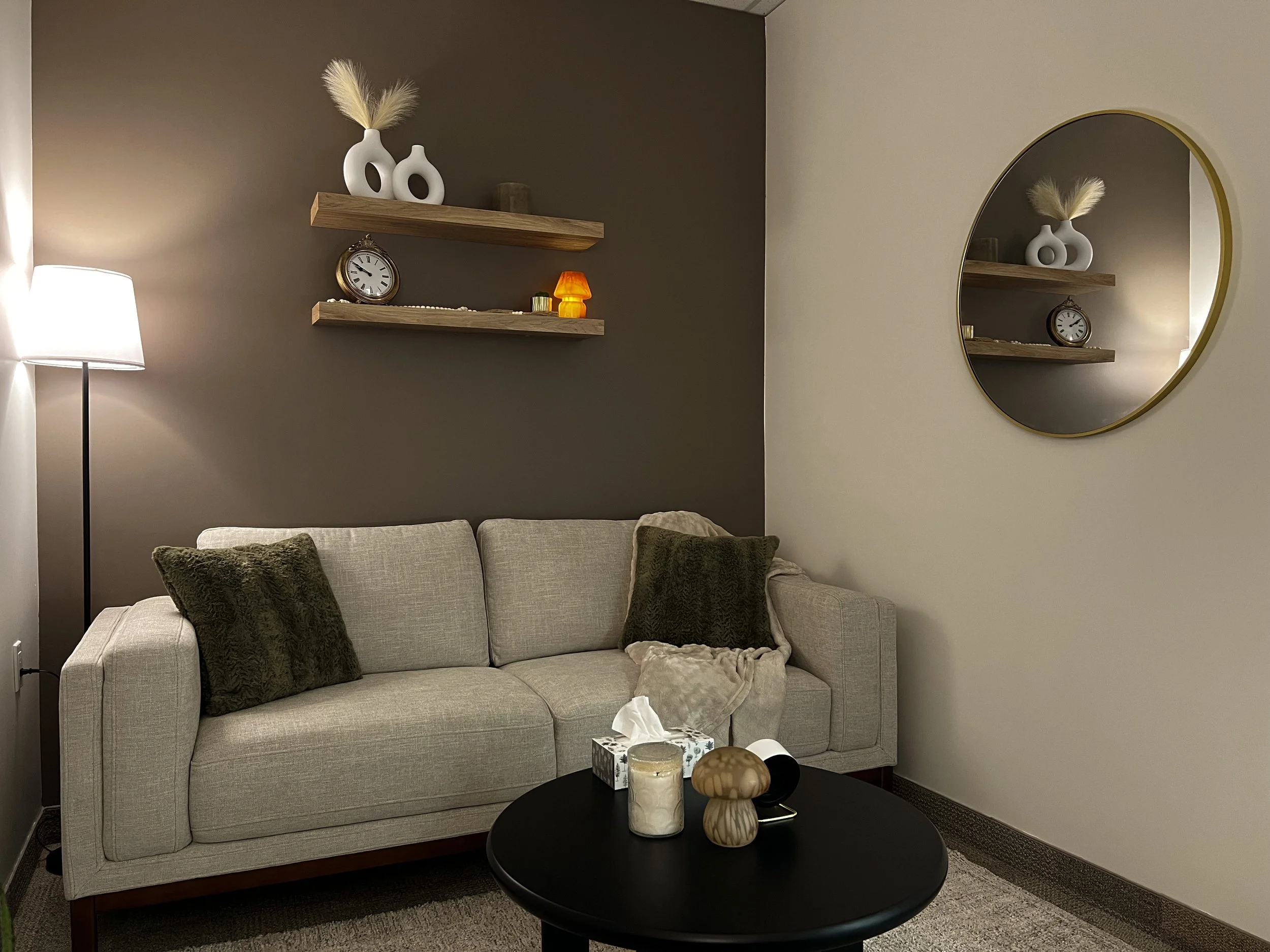 Living room with beige sofa, dark pillows, beige blanket, and round black coffee table containing candles and decorative objects. Wall behind upholstered in dark gray with wooden shelves holding vases, clock, and decor. Round mirror on adjacent wall reflecting the scene, beside a floor lamp.