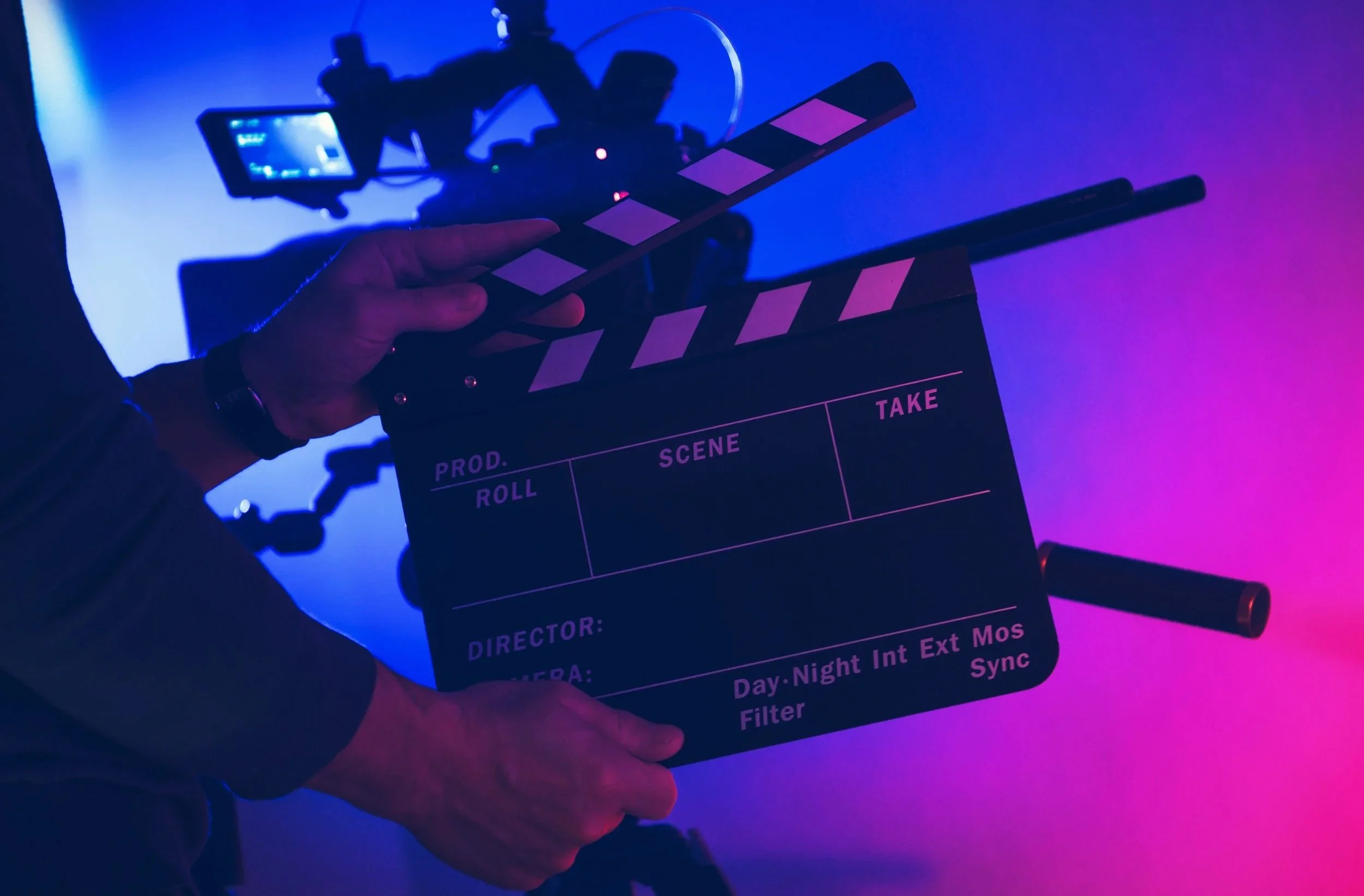 Close-up of a person's hand holding a black and white film clapperboard in a studio with colorful blue and pink lighting, with a camera mounted on a rig in the background.