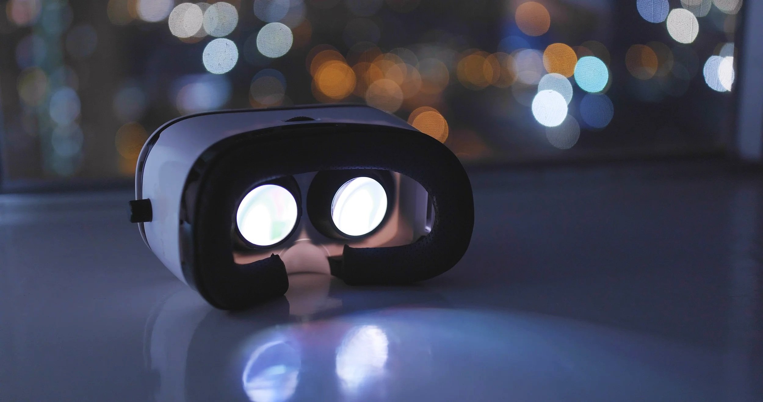 A virtual reality headset resting on a surface with a blurred cityscape with colorful lights in the background.