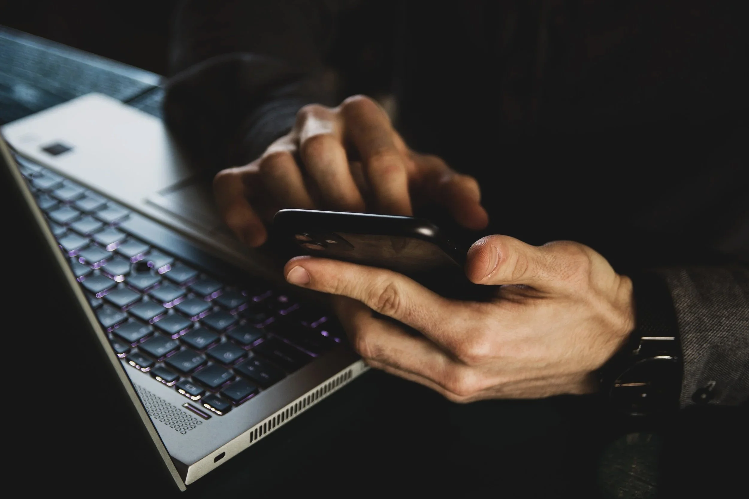 Close-up of a person with a watch on their wrist, using a smartphone with a laptop computer nearby.