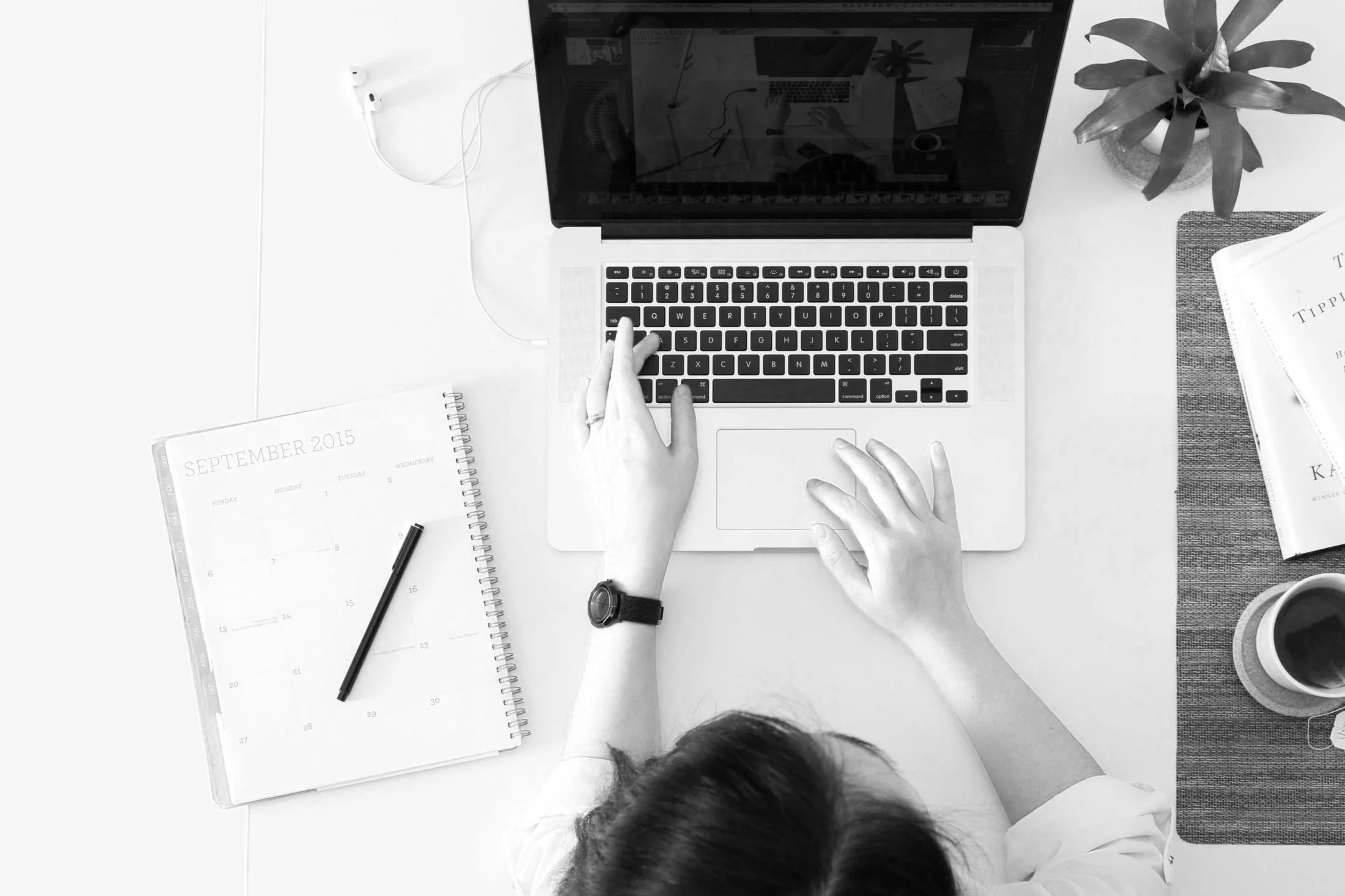Top-down view of a person working on a laptop at a desk with a planner, a pen, a book, a cup of coffee, and a plant.