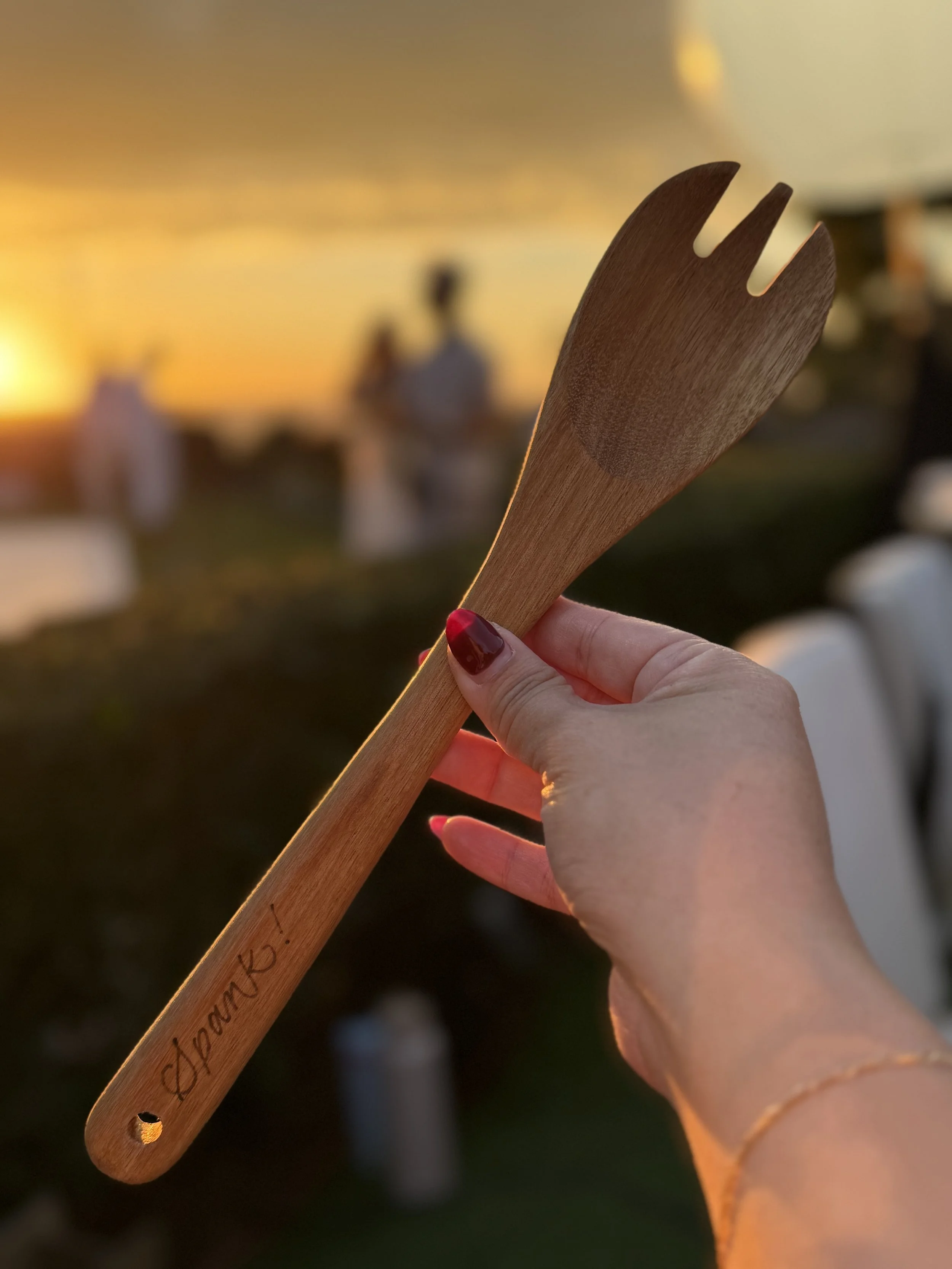 A hand holding a wooden fork with a sunset in the background, with blurred figures and chairs in an outdoor setting.