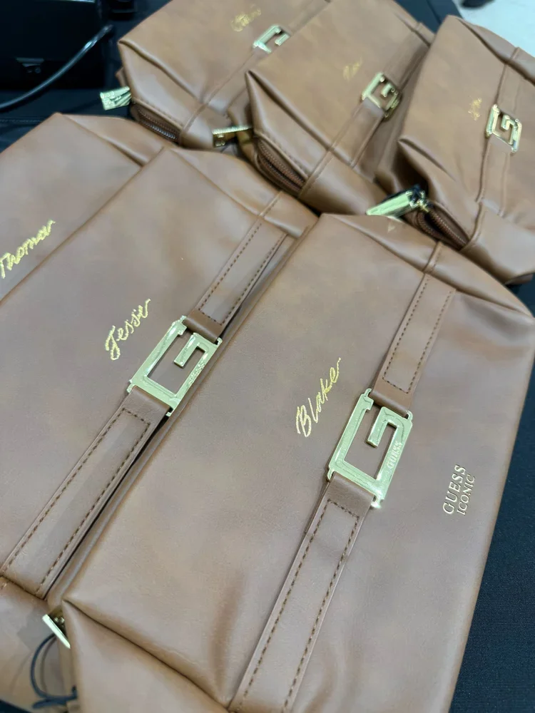 Four brown backpacks with gold embroidered names and gold hardware, labeled with personal names and the brand 'Guess Iconic'.