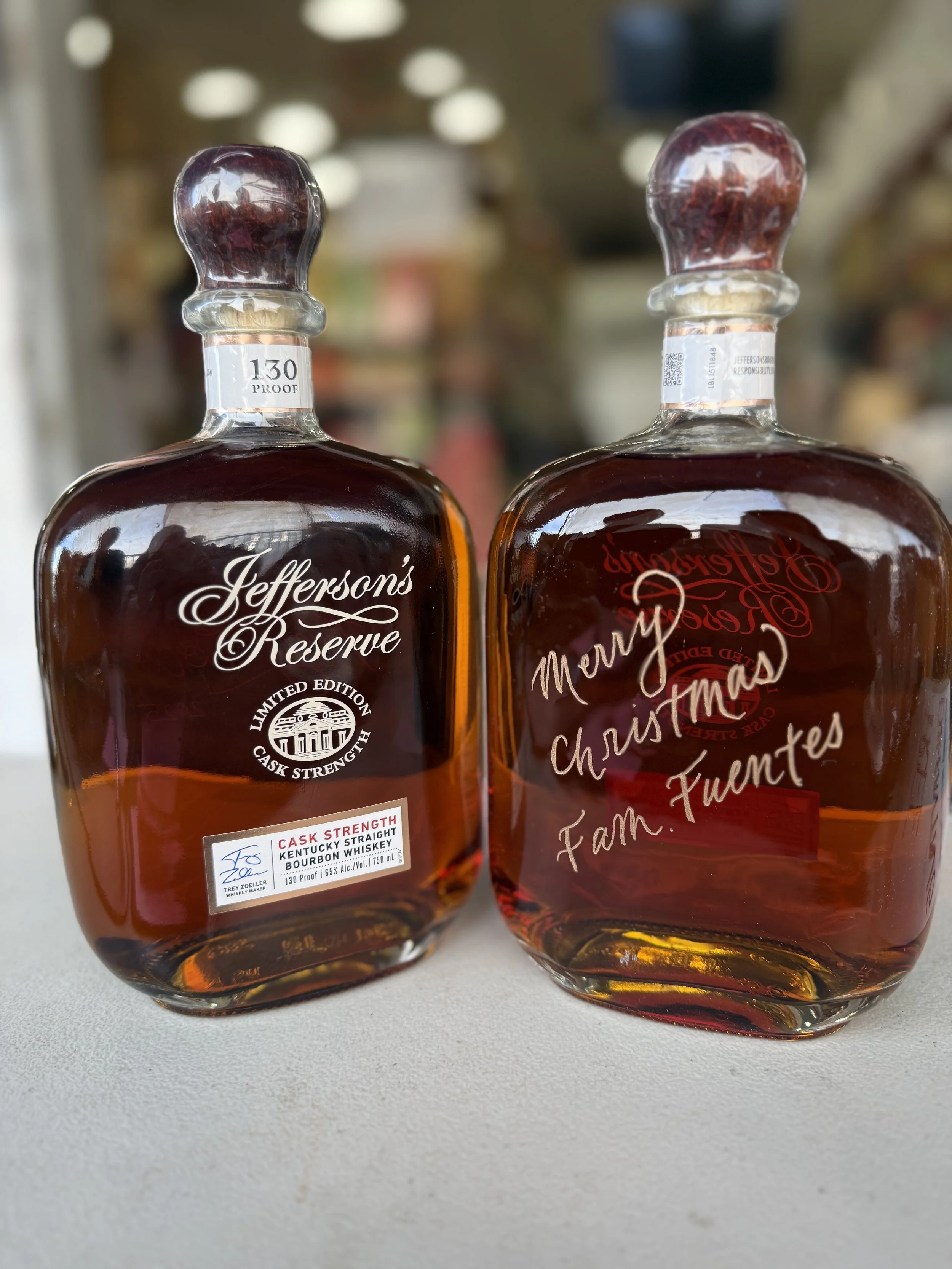 Two bottles of Jefferson's Reserve bourbon whiskey on a light surface, with a blurred store background. The bottle on the left is labeled as Cask Strength Kentucky Straight Bourbon Whiskey, 130 proof. The bottle on the right has a holiday message tha