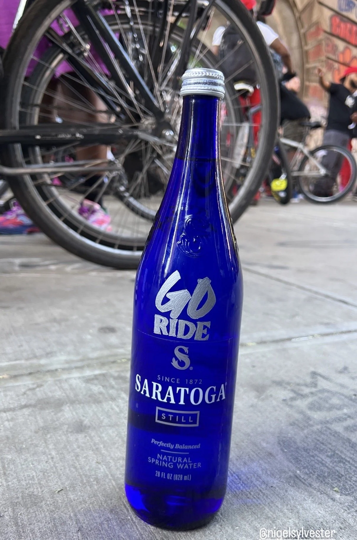 Blue glass bottle of Saratoga Still Natural Spring Water placed on a sidewalk, with bicycles and people in the background.