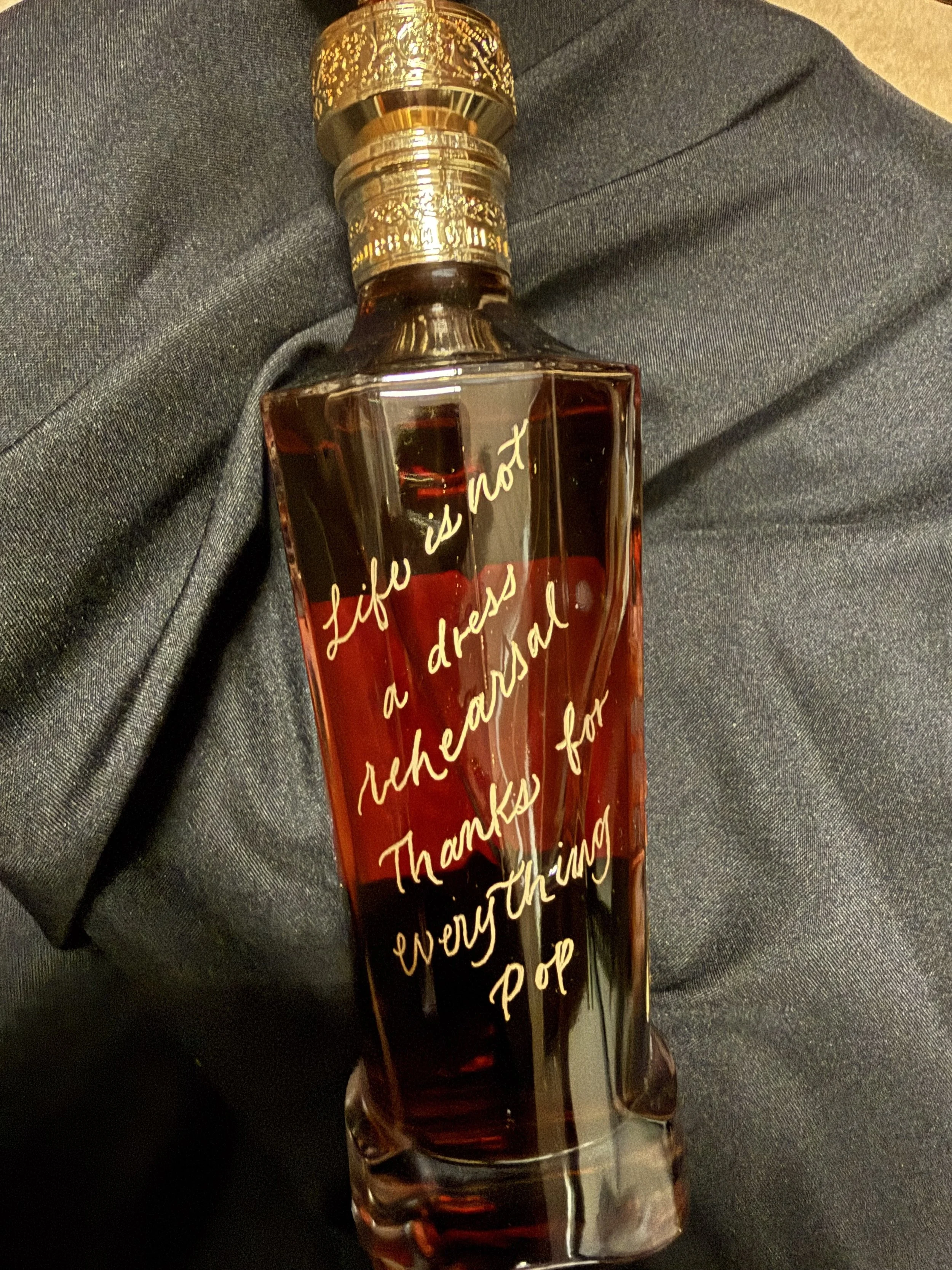 A decorative perfume or cologne bottle made of clear glass with dark liquid inside. The bottle has a gold-colored cap and features cursive gold-writing that says, 'Life is not a dress rehearsal. Thanks for everything. P.O.P.'