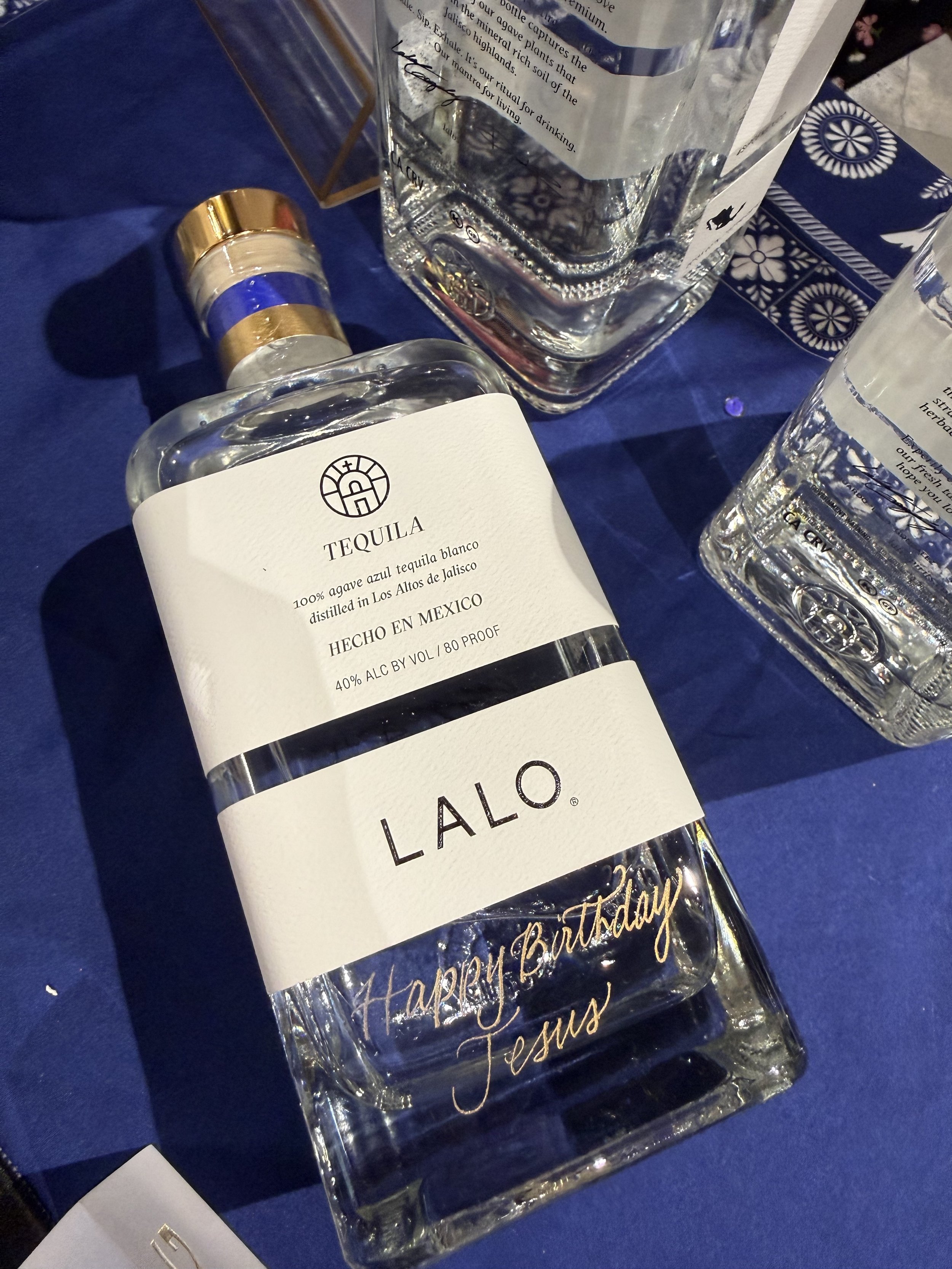 A bottle of Lalo tequila with a personalized 'Happy Birthday Jesus' message on the front, placed on a blue tablecloth alongside other bottles and glasses.