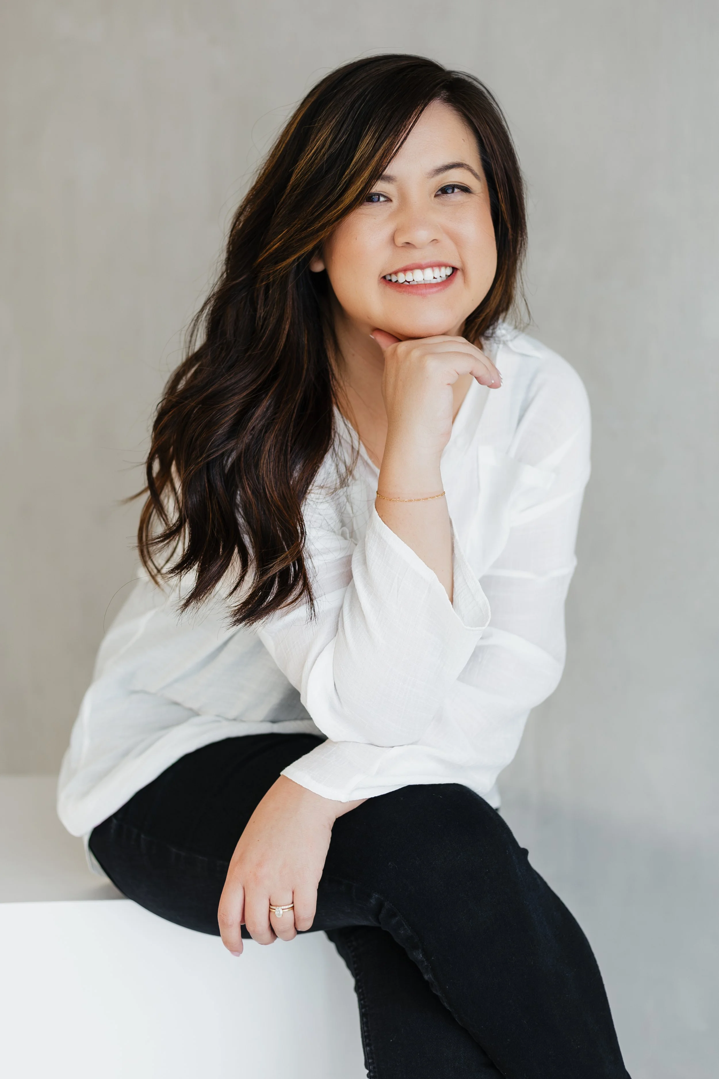 A female Los Angeles calligrapher with long dark brown hair, smiling and sitting on a white surface, wearing a white blouse and black pants.