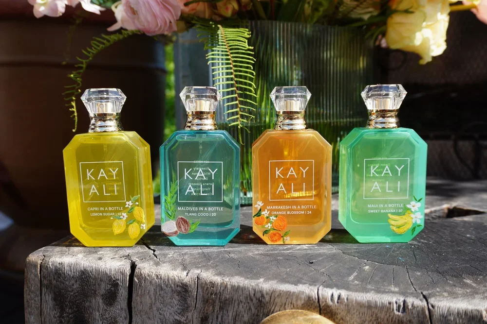 Four colorful perfume bottles with clear, faceted caps on a weathered wooden surface, surrounded by green plants and flowers in the background. Each bottle has a label with the brand 'Kayali' and a fragrance name.
